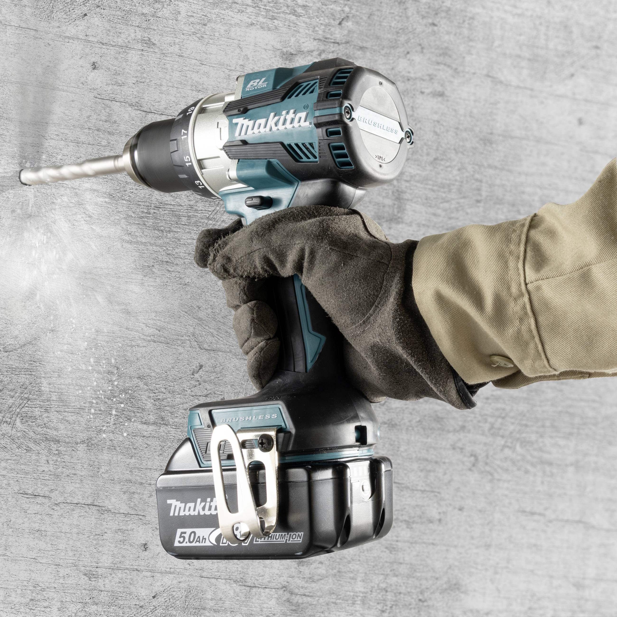 Person mit Handschuh hält eine elektrische Bohrmaschine an einer Betonwand. Die Bohrmaschine ist in Gebrauch, Staub entsteht.