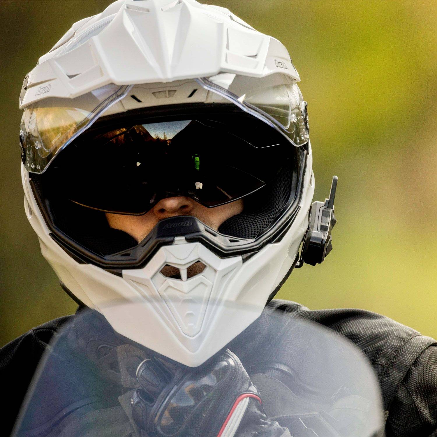 Eine Person trägt einen weißen Motorradhelm und eine schwarze Schutzbrille, im Hintergrund ist unscharfes Grün und Blau zu sehen.
