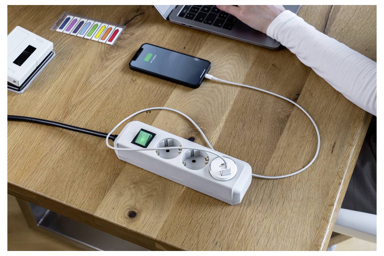 Smartphone wird von einer Steckerleiste neben einem Laptop auf einem Holzschreibtisch aufgeladen, während eine Person in der Nähe arbeitet und Bürogegenstände im Hintergrund sichtbar sind.