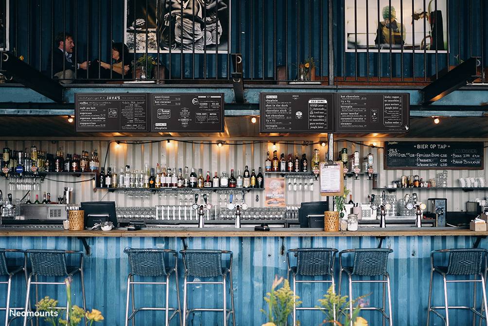 Eine moderne Bar im Freien mit blauen Metallwänden. Oben sind Tafeln mit Getränkemenüs, darunter eine Theke mit Barhockern.