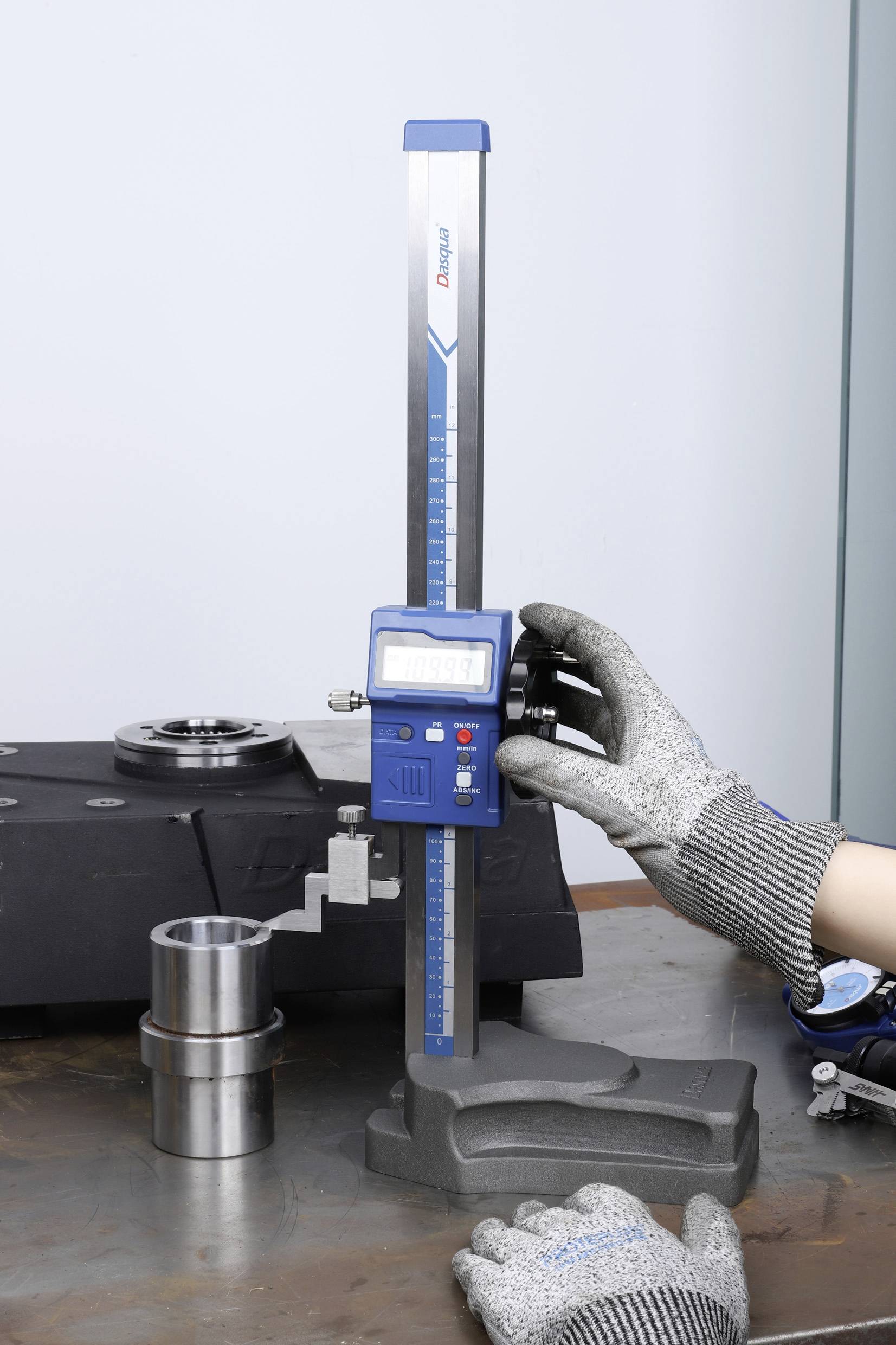 Eine Person misst ein Metallobjekt auf einem Tisch mit einer digitalen Höhenmesslehre. Trägt Arbeitshandschuhe.