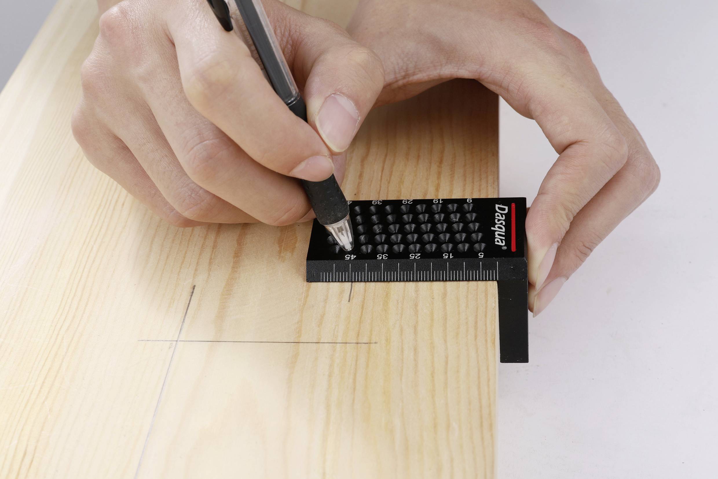 Eine Hand hält einen Stift und markiert mit einem schwarzen Lineal präzise auf einem Holzbrett eine Linie für Messzwecke.