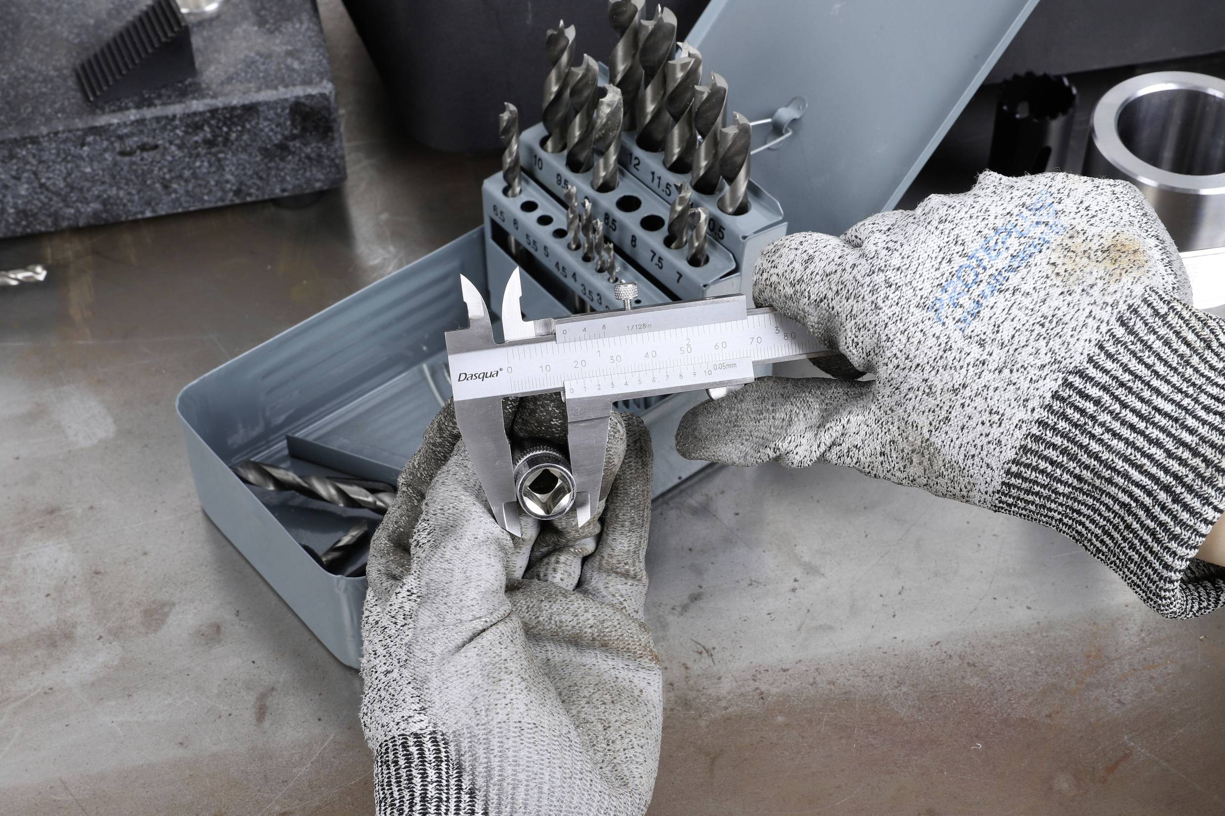 Person mit grauen Handschuhen hält eine Messschieber und misst einen Metallgegenstand, während daneben Metallbohrer in einer Box liegen.