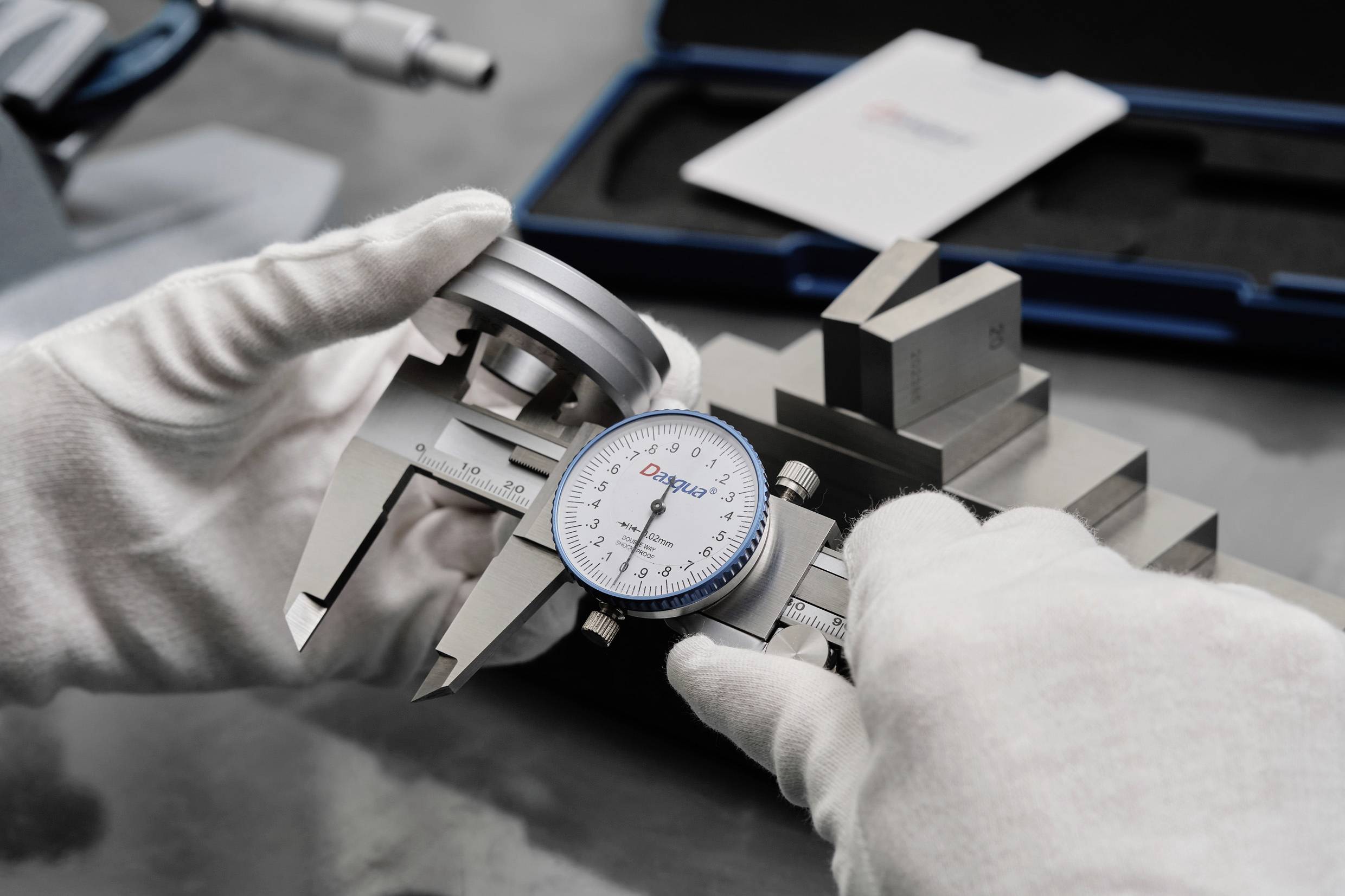 Eine Person misst ein Metallstück mit einem analogen Messschieber. Handschuhe schützen vor Fingerabdrücken. Im Hintergrund sind Werkzeuge erkennbar.