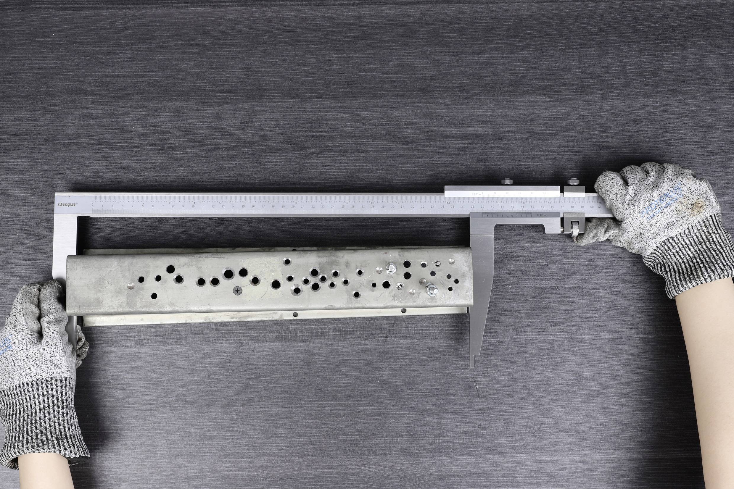 Eine Person misst ein rechteckiges Metallteil mit einer Schieblehre. Das Teil hat mehrere Löcher unterschiedlicher Größe.