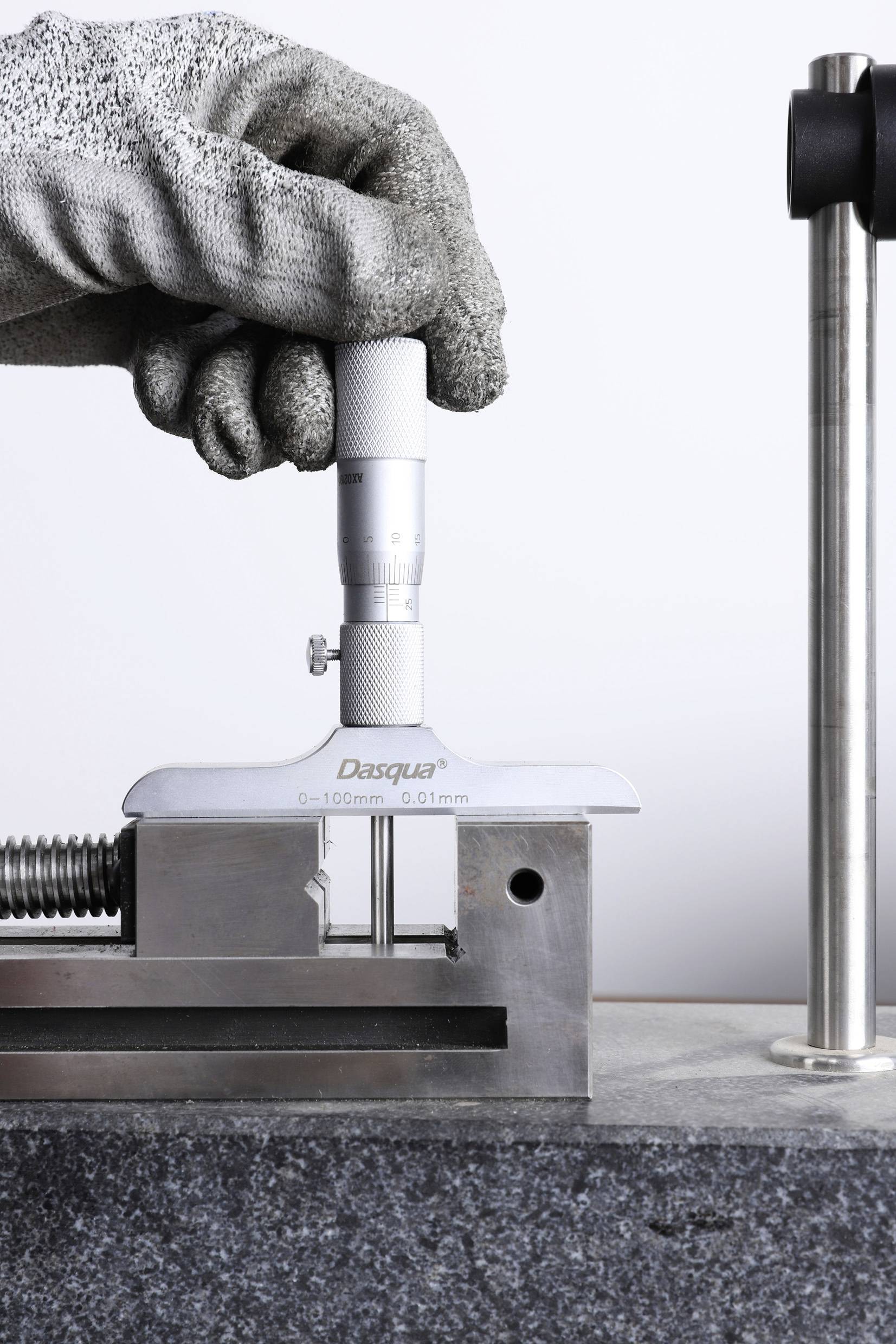 Eine Hand in einem Handschuh hält ein Höhenmessgerät, das ein Metallstück misst, montiert auf einer Werkbank.