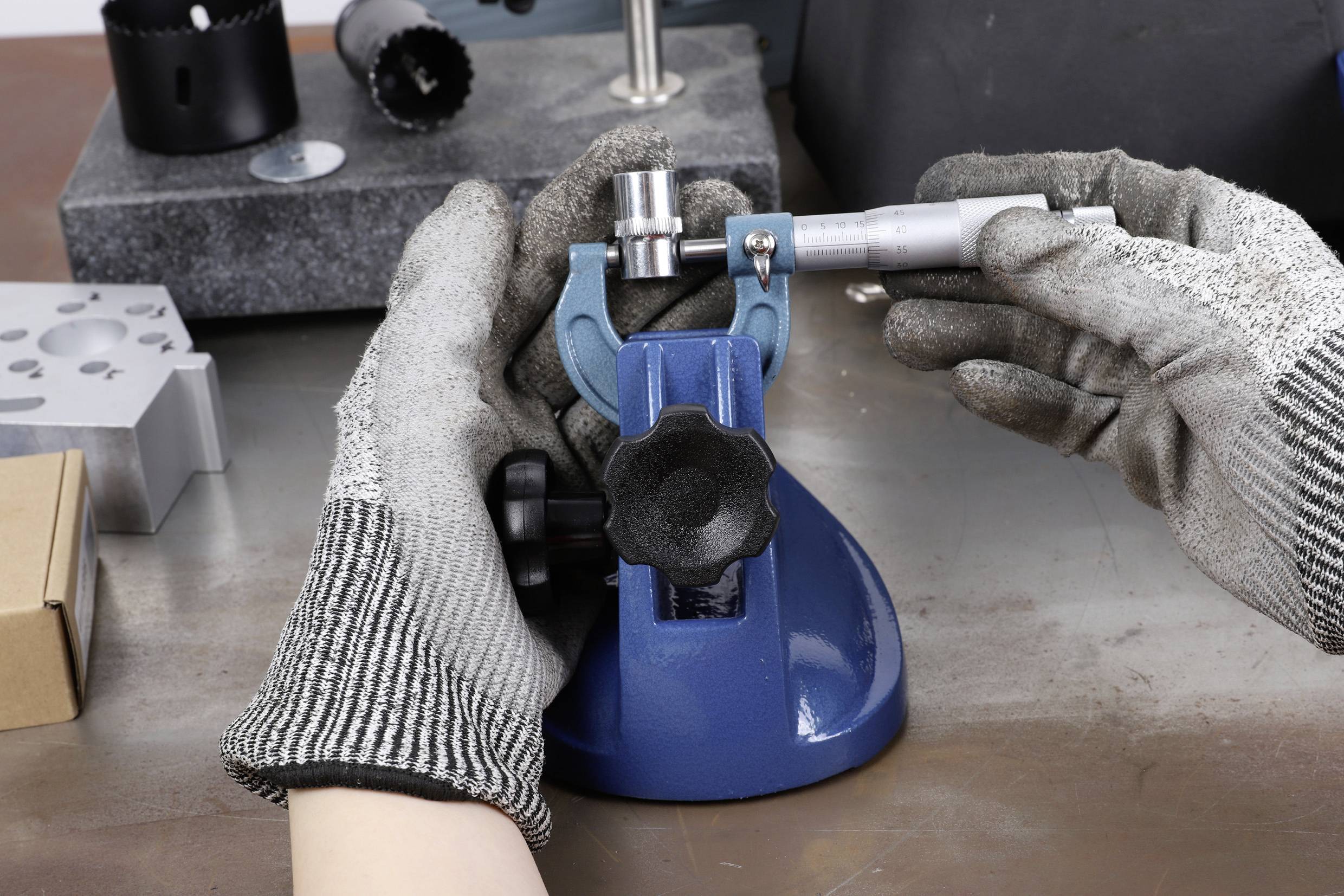 Eine Person trägt Handschuhe und bedient ein blaues Präzisionsmessgerät auf einem Arbeitstisch in einer Werkstattumgebung.