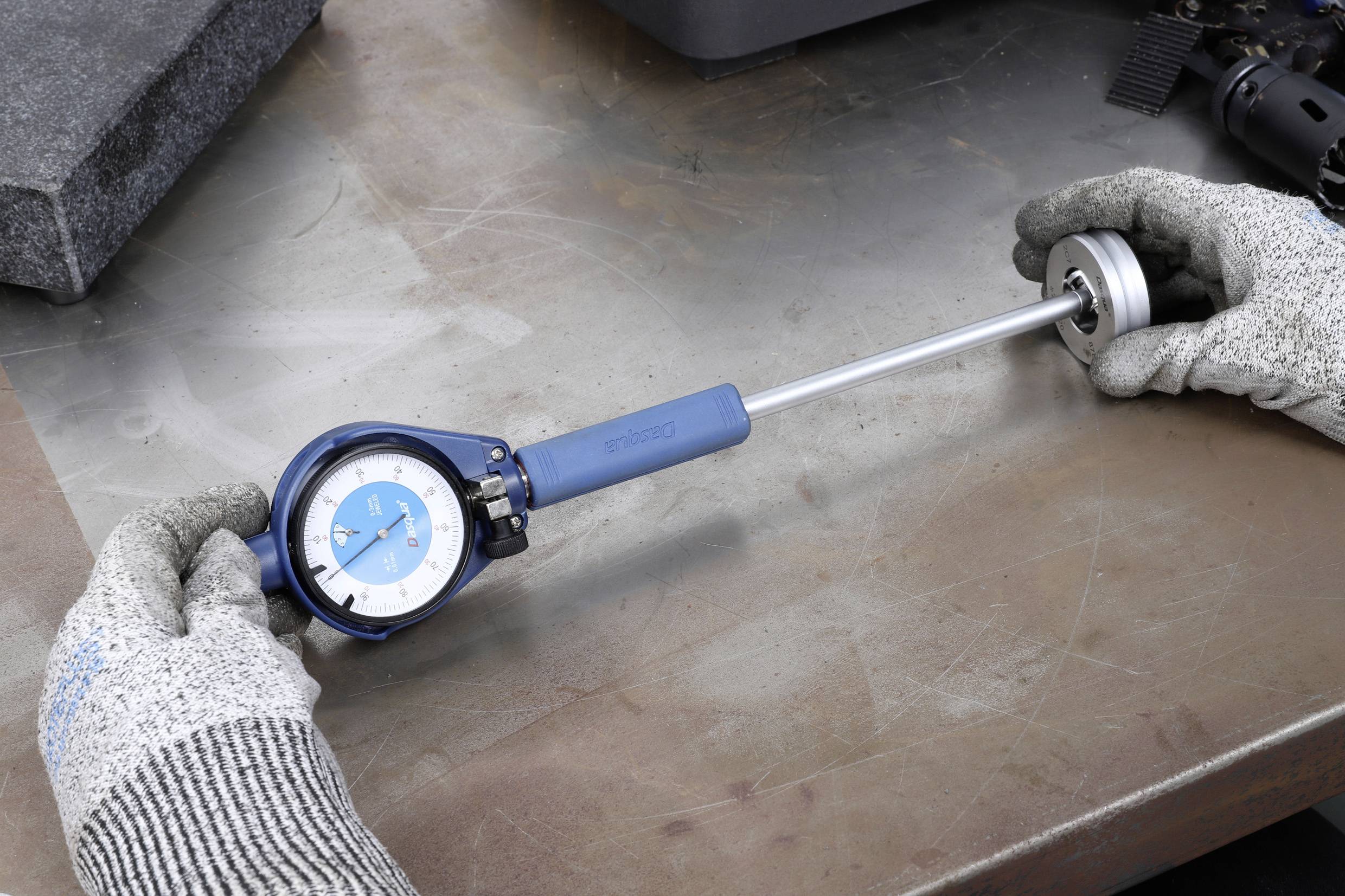 Zwei Hände in Handschuhen halten ein Werkzeug zur Messung des Innendurchmessers von zylindrischen Objekten auf einem Metalltisch.