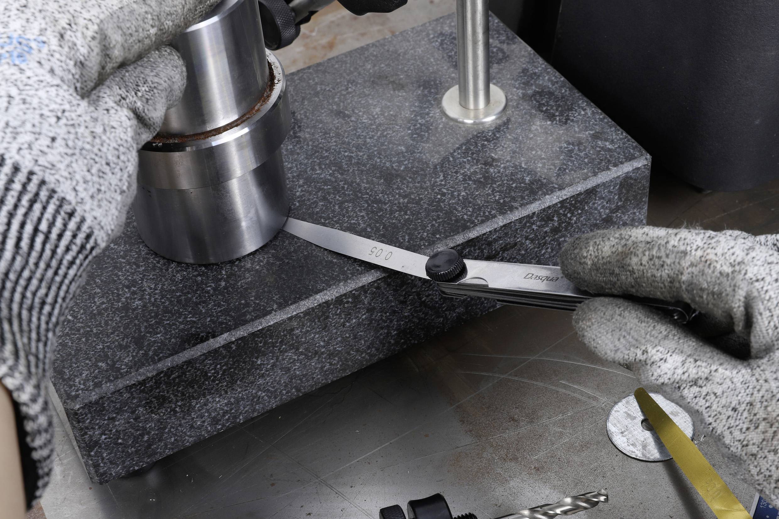 Zwei behandschuhte Hände messen mit einem Messinstrument den Durchmesser eines Metallzylinders, der auf einer Granitplatte steht.