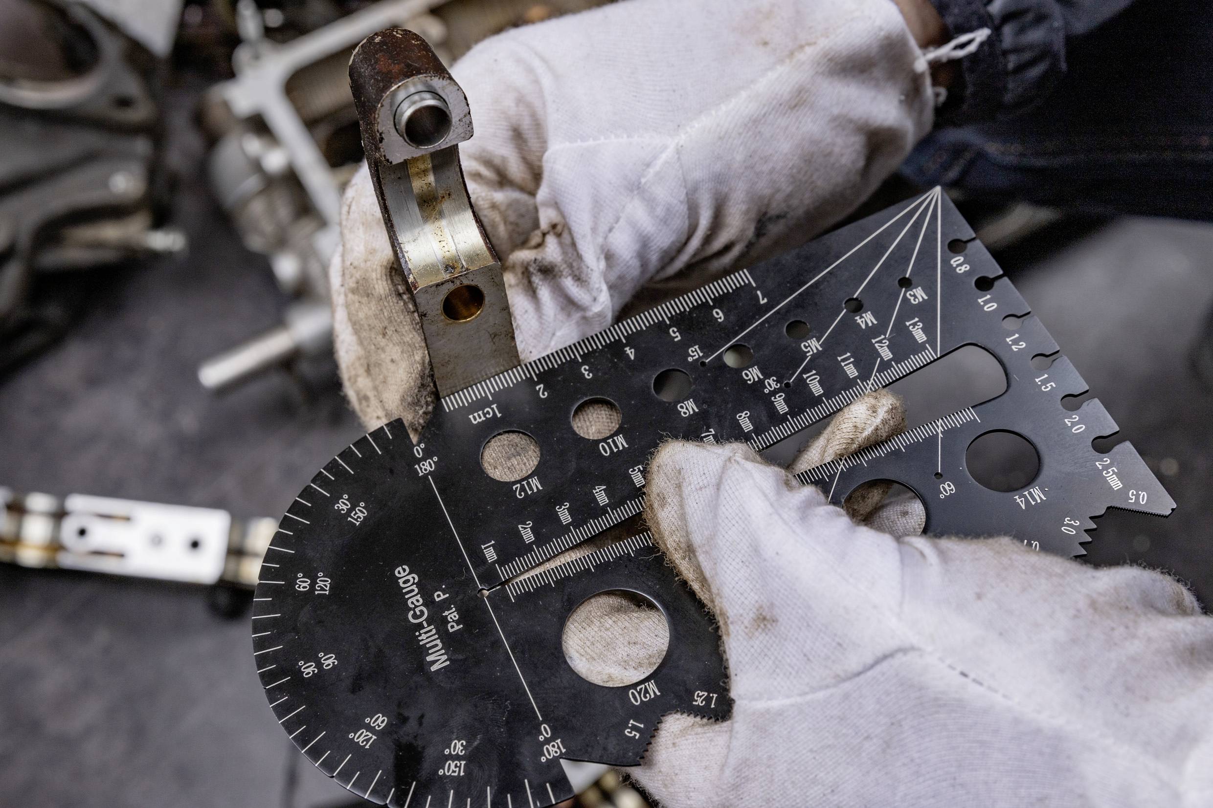Zwei behandschuhte Hände halten ein Winkelmessgerät, um die Größe eines Metallteils zu überprüfen. Im Hintergrund liegen Werkzeuge.