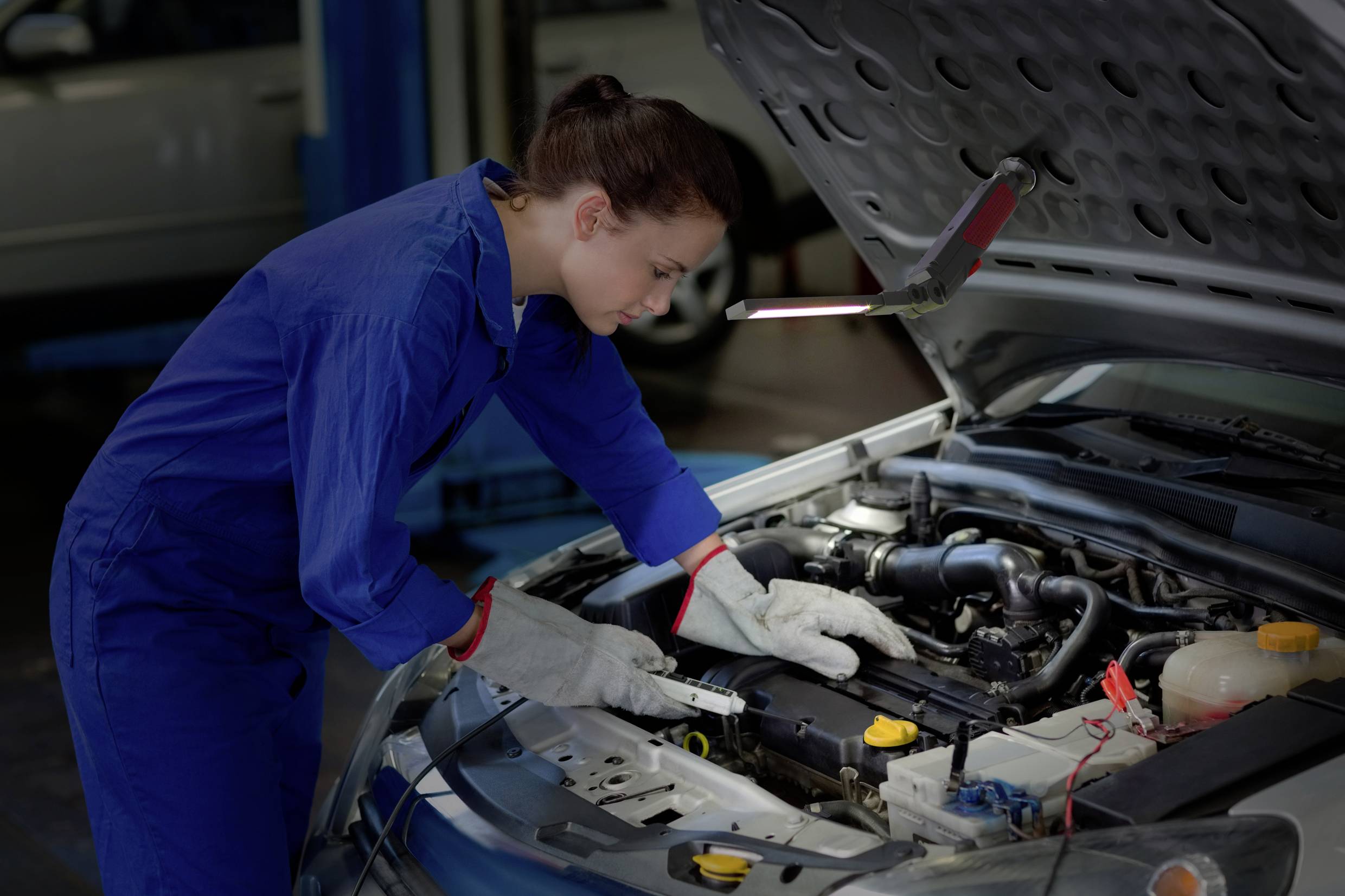Eine Frau in blauer Arbeitskleidung repariert unter der Motorhaube eines Autos in einer Werkstatt.