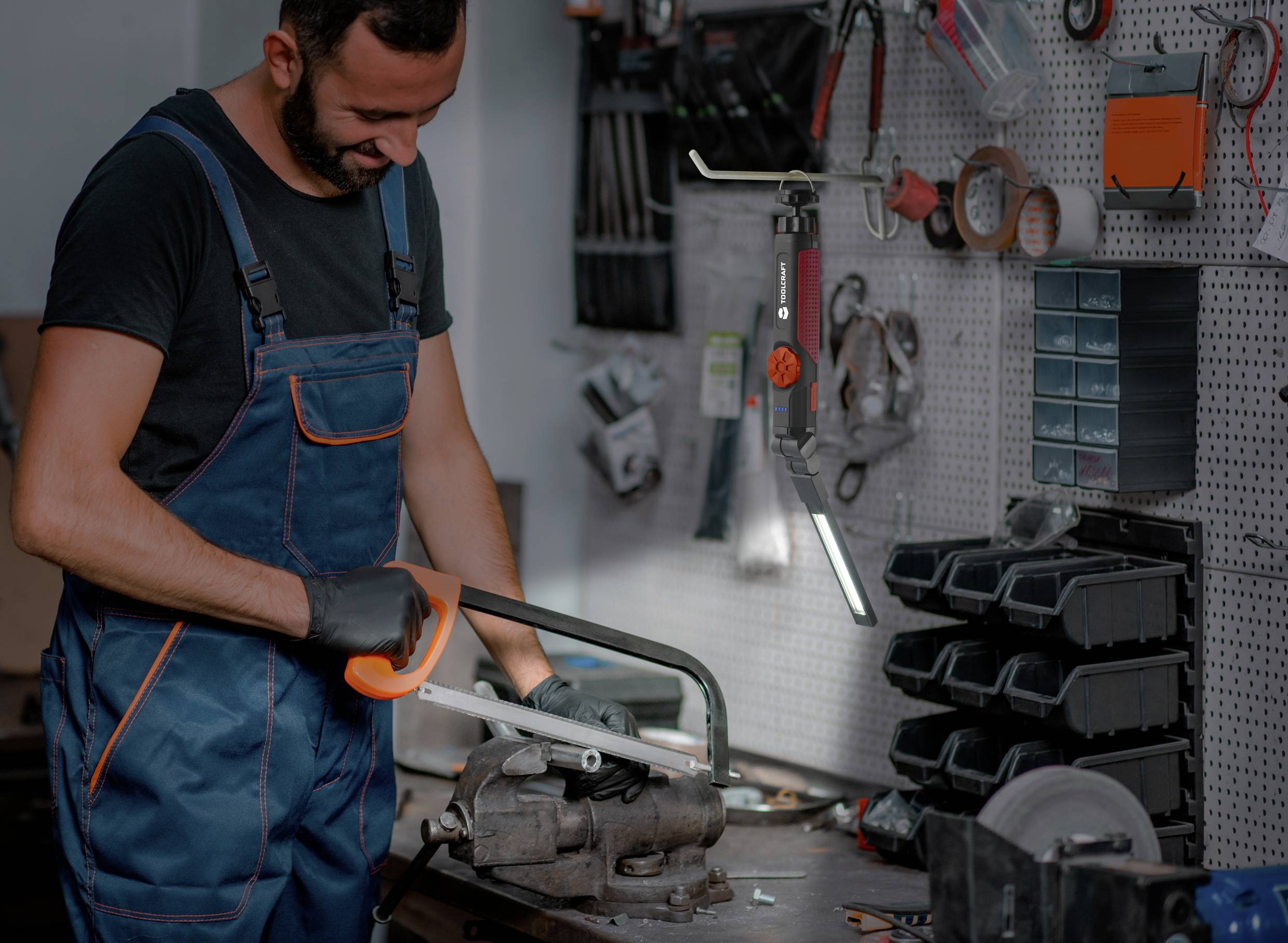 Ein Mann in Arbeitskleidung sägt in einer Werkstatt mit einer Handsäge durch ein Metallrohr. Werkzeuge hängen an der Wand.