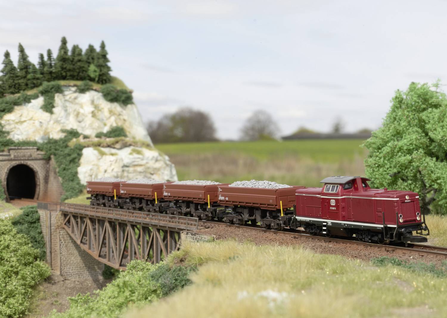 Ein roter Modellzug mit offenen Waggons fährt über eine Brücke in einer Landschaft mit Bäumen, einem Tunnel und einem Hügel.
