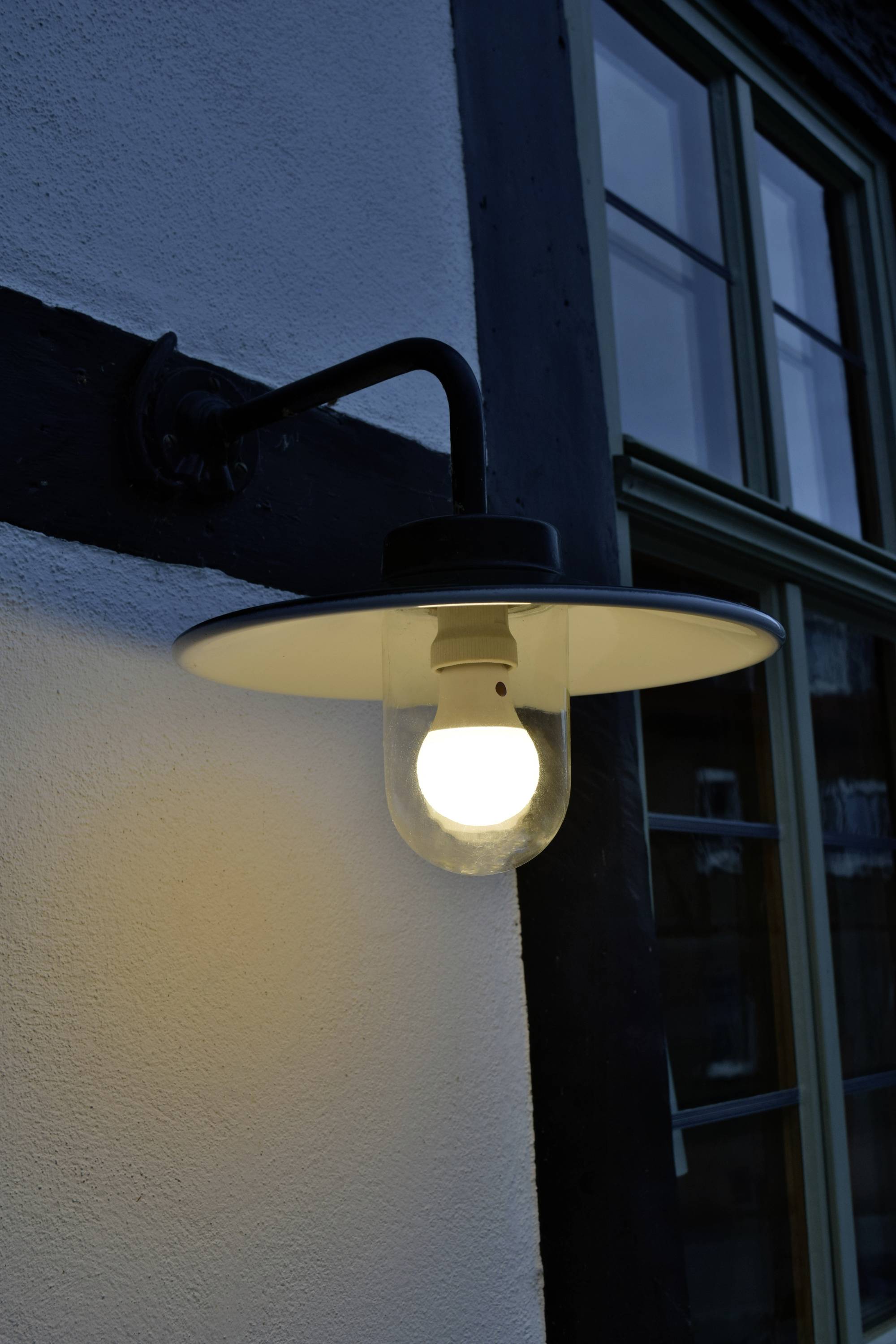 Eine eingeschaltete Außenlampe an einer weißen Wand neben einem Fenster, beleuchtet die Umgebung in der Dämmerung.