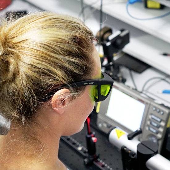 Eine Frau mit Schutzbrille arbeitet an einem wissenschaftlichen Laborinstrument. Im Hintergrund ist ein Bildschirm sichtbar.