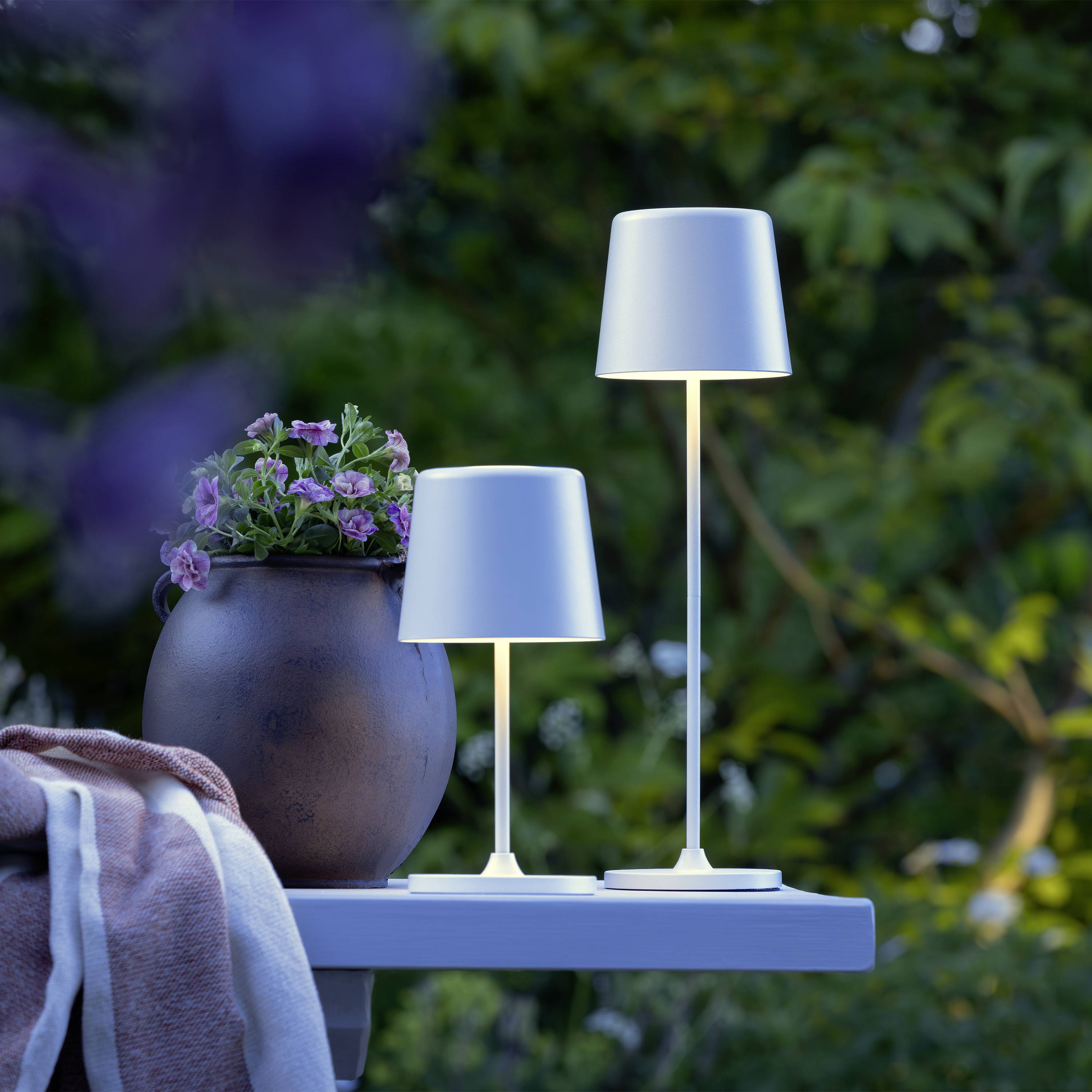 Zwei moderne Tischlampen und ein Blumentopf mit lila Blumen stehen auf einem Gartentisch im Freien vor grünem, unscharfem Hintergrund.