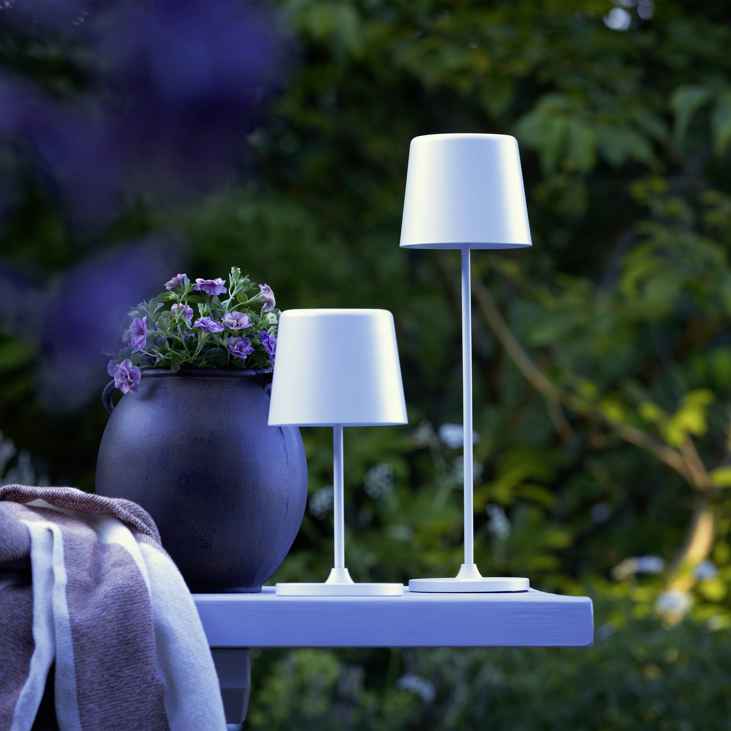 Ein gedeckter Gartentisch im Freien mit zwei schlanken, modernen Tischlampen und einem Topf mit lila Blumen, umgeben von Pflanzen.