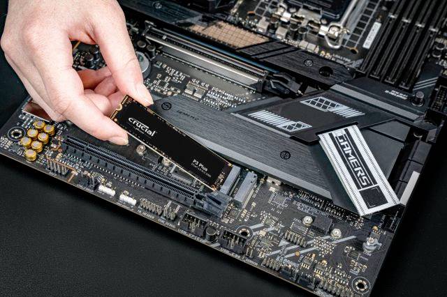Eine Hand installiert eine SSD auf einem Motherboard. Der Steckplatz und andere elektronische Komponenten sind sichtbar.