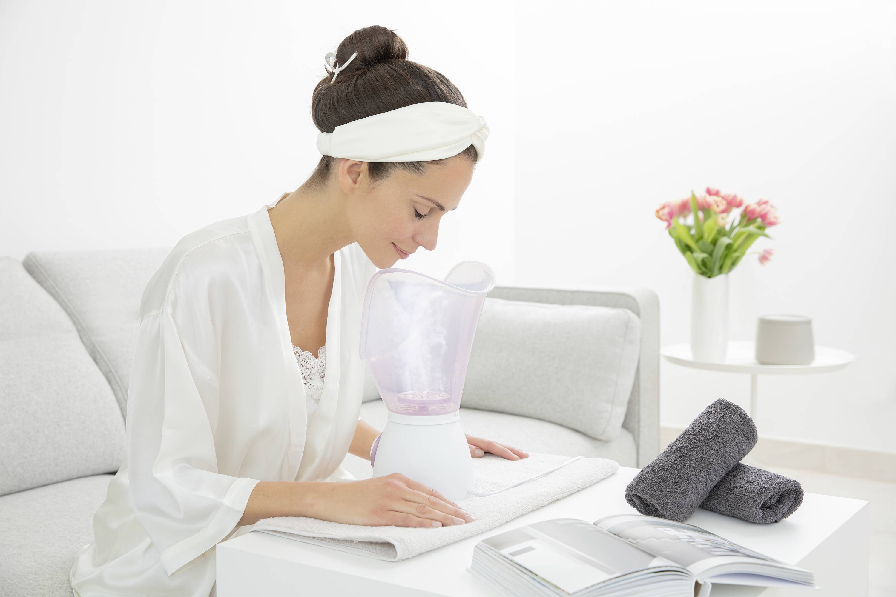 Eine Frau sitzt auf der Couch und nutzt einen Gesichtsdampfer. Neben ihr liegen Handtücher und ein offenes Buch. Im Hintergrund steht eine Vase mit Blumen.