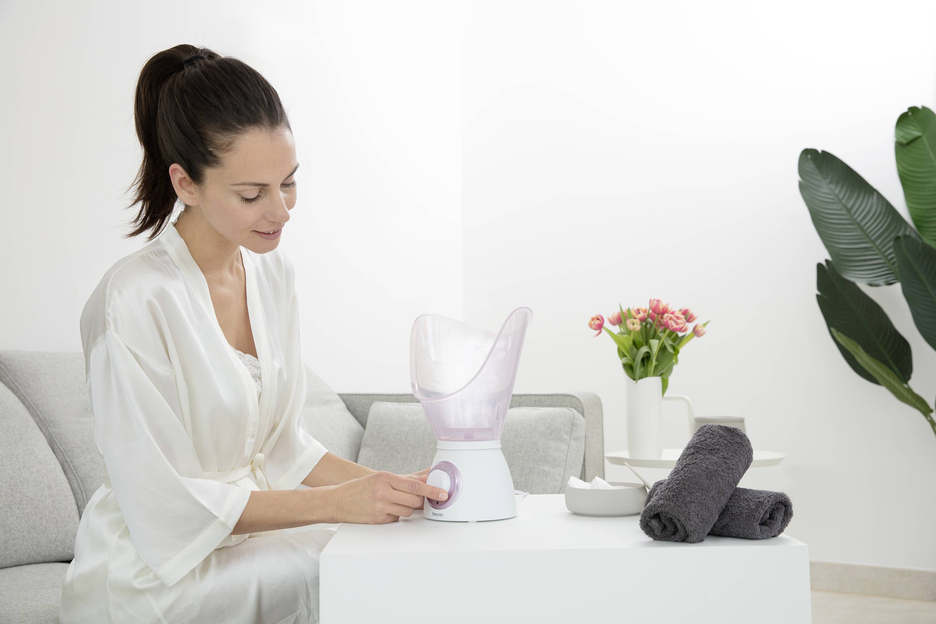 Frau sitzt auf dem Sofa und verwendet einen Gesichtsdampfer. Neben ihr liegen Handtücher und eine Vase mit Blumen.