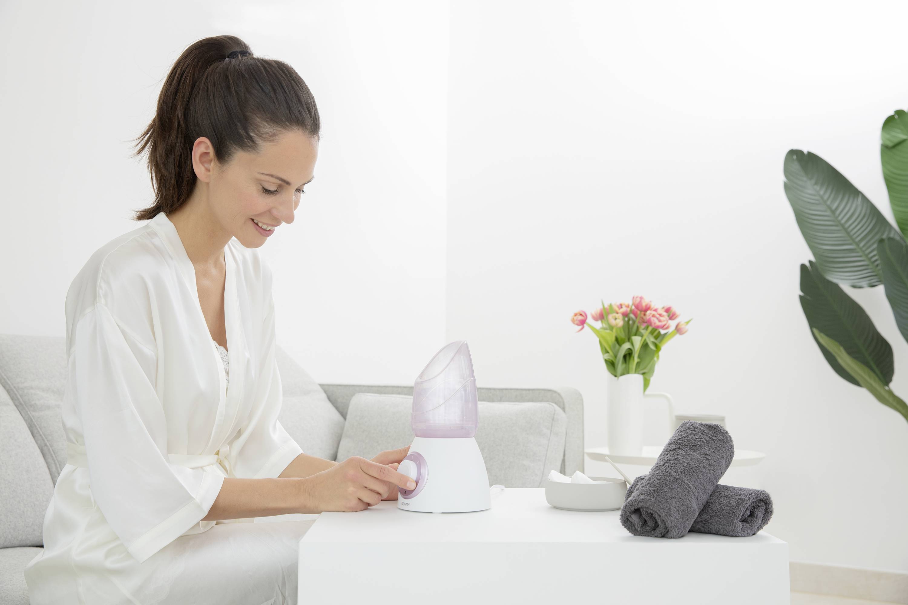 Eine Frau benutzt einen Gesichtsdampfer in einem hellen Wohnzimmer. Neben ihr sind Handtücher und Blumen auf einem Tisch arrangiert.