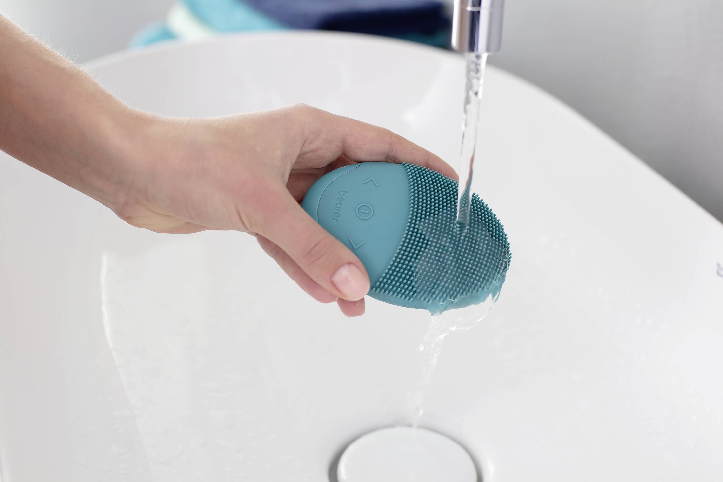 Hand hält ein blaues Gesichtsreinigungsgerät unter fließendem Wasser über einem Waschbecken.