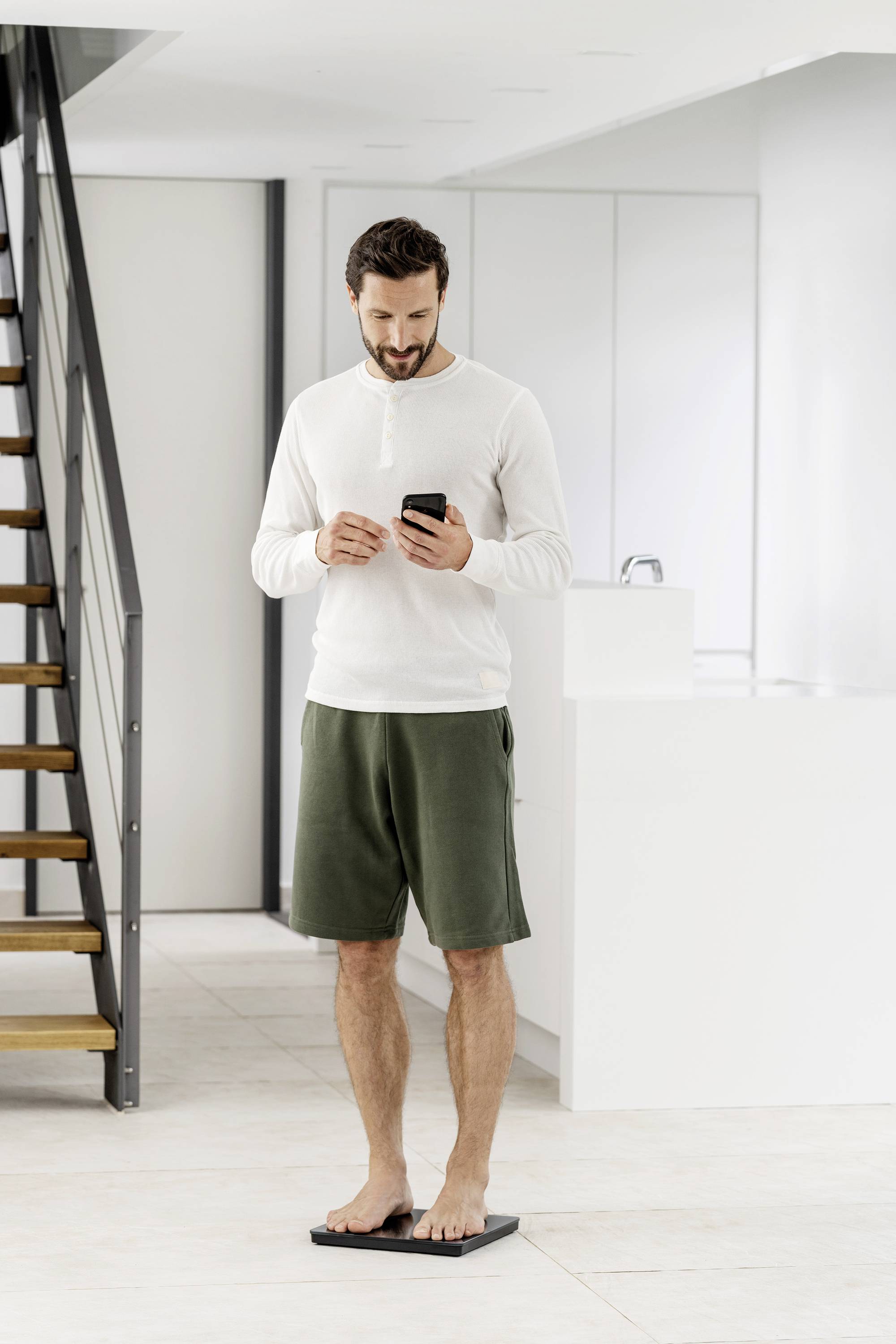 Ein Mann steht auf einer Waage in einem modernen, hellen Raum und schaut auf sein Smartphone.
