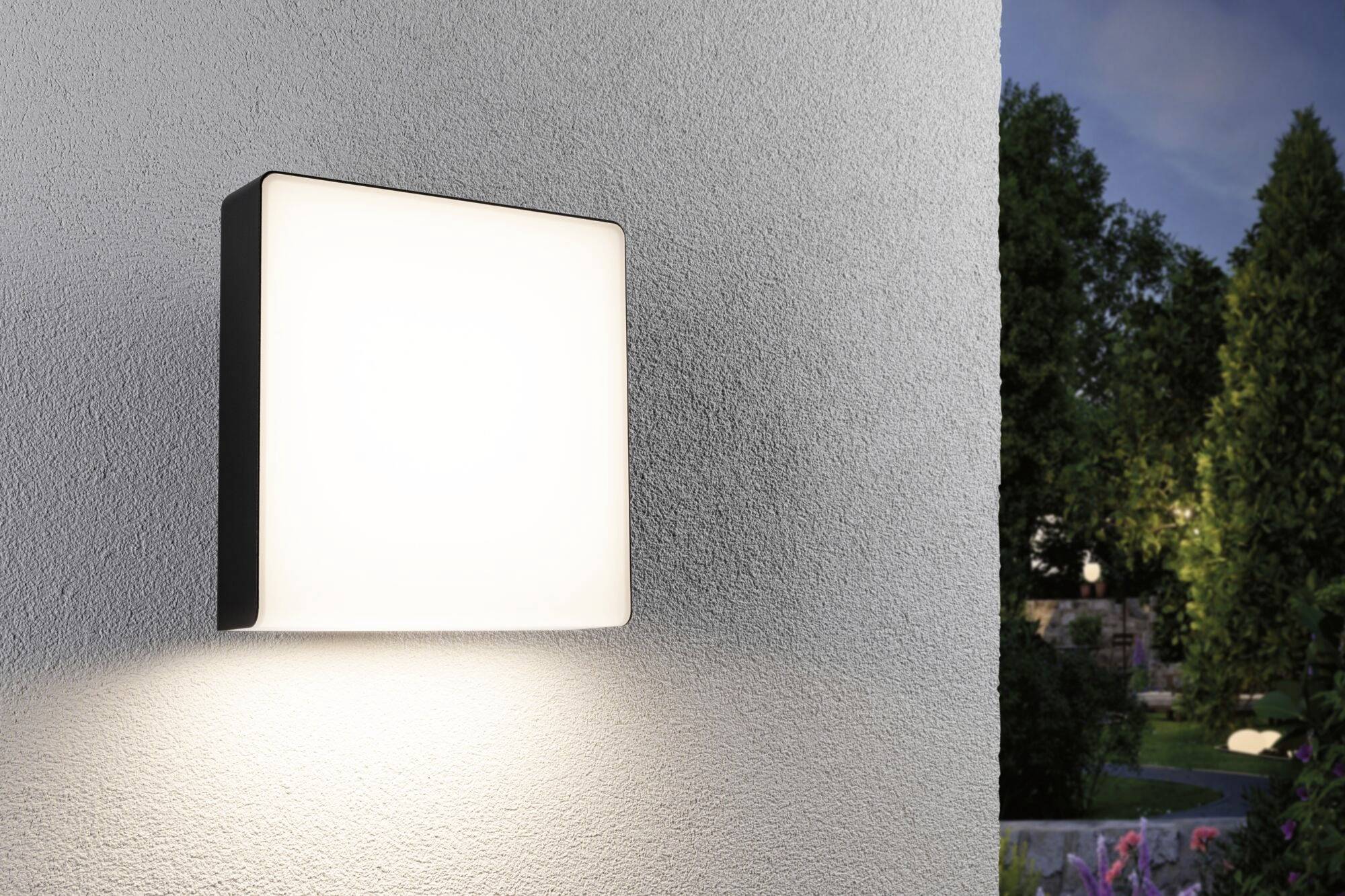 Außenleuchte an einer weißen Wand leuchtet bei Dämmerung, beleuchtet die Umgebung neben einem Gartenbereich.