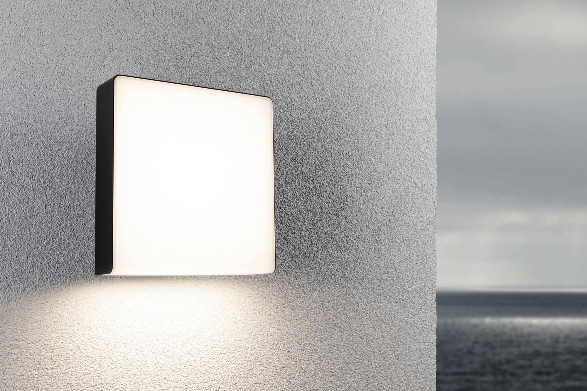 Wandleuchte an einer Wand, beleuchtet einen Bereich im Freien mit Meerblick im Hintergrund.