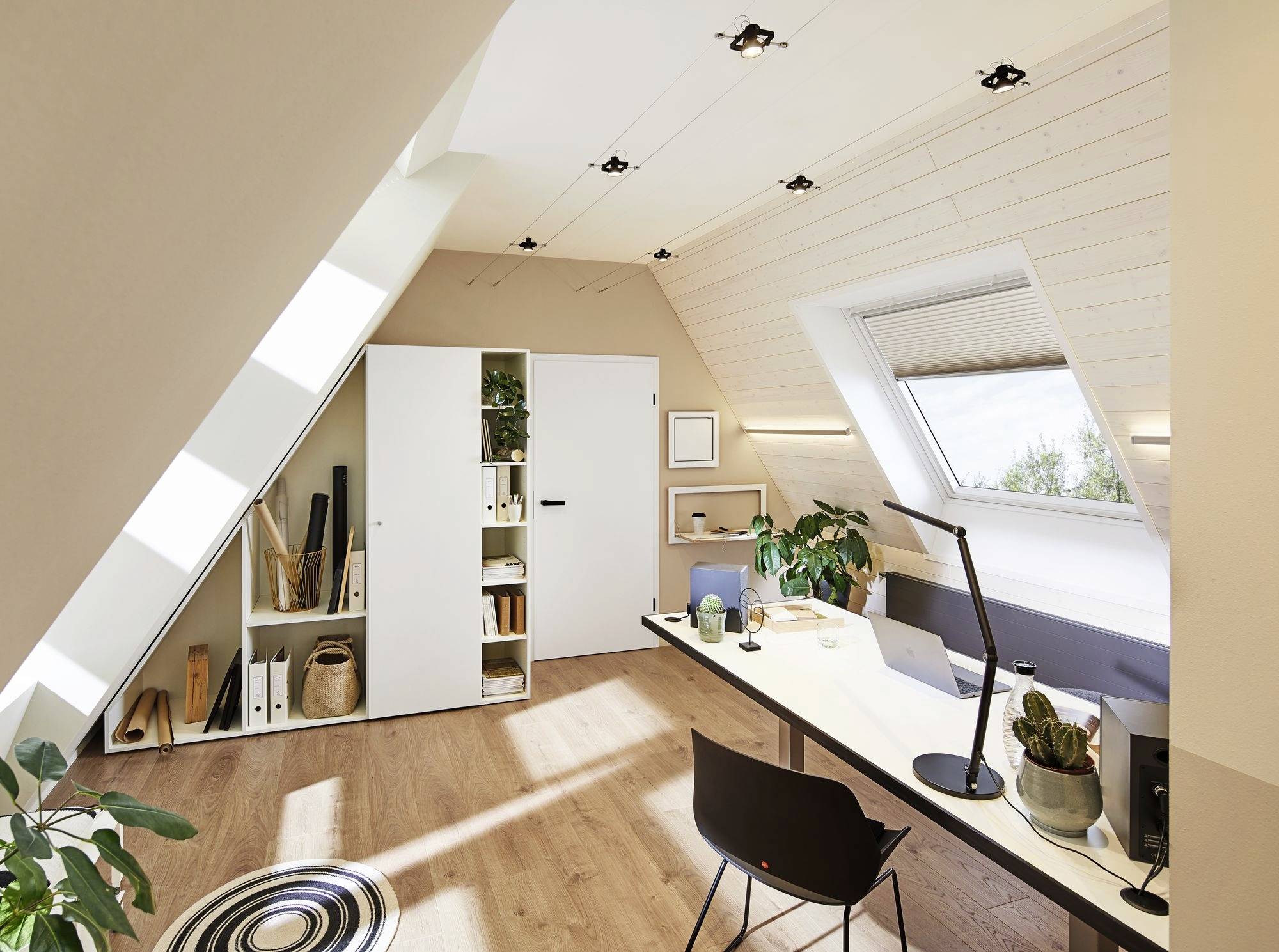 Helles Dachgeschossbüro mit Holzfußboden, Schreibtisch, Stuhl, Pflanzen und Regalen im Hintergrund. Große Fenster lassen viel Licht herein.