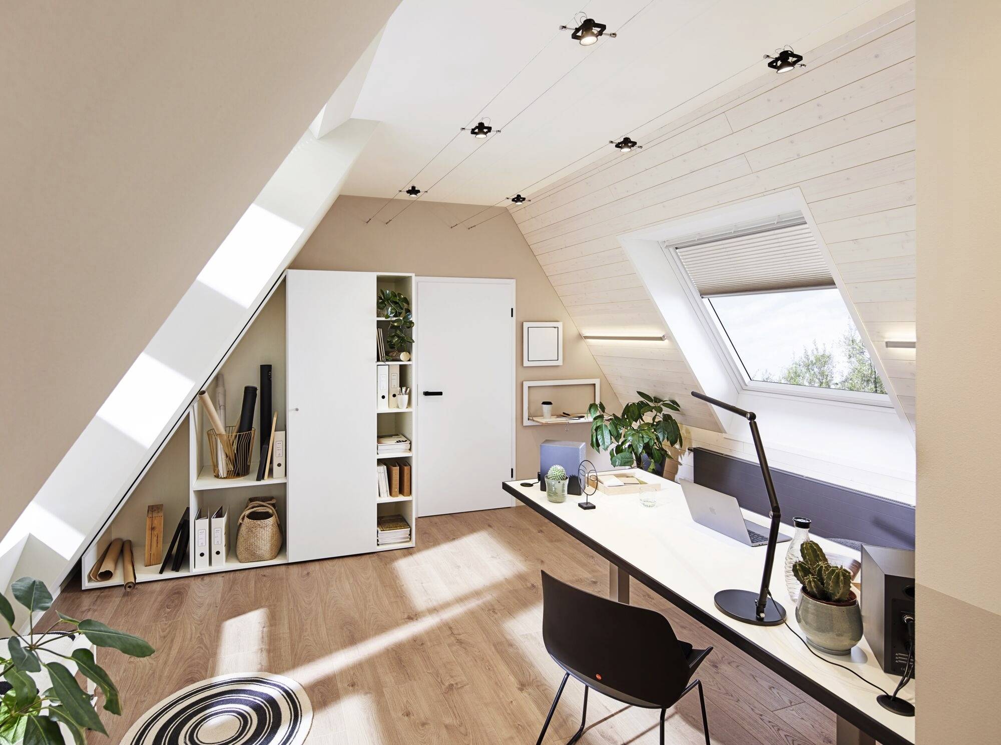 Helles Homeoffice in einem Dachgeschoss mit weißem Schreibtisch, schwarzem Stuhl, Pflanzen und Regalen, durch Dachfenster beleuchtet.