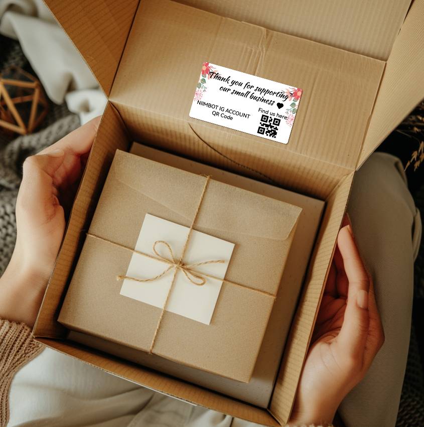 Person hält einen geöffneten Karton mit einem kleineren Geschenkpaket darin. Aufkleber außen trägt den Text 'Thank you for supporting our small business'.