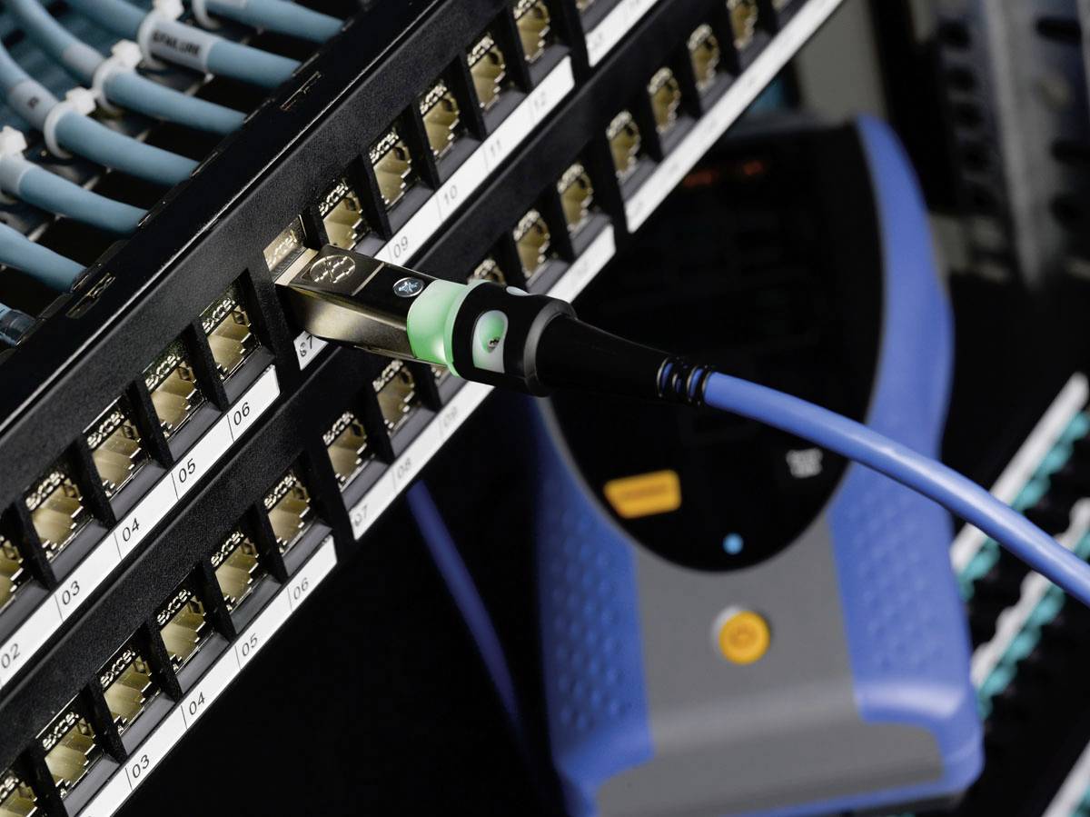 Ein Netzwerkkabel ist in einen nummerierten Port eines Server-Racks eingesteckt. Im Hintergrund ist ein Testgerät zu sehen.