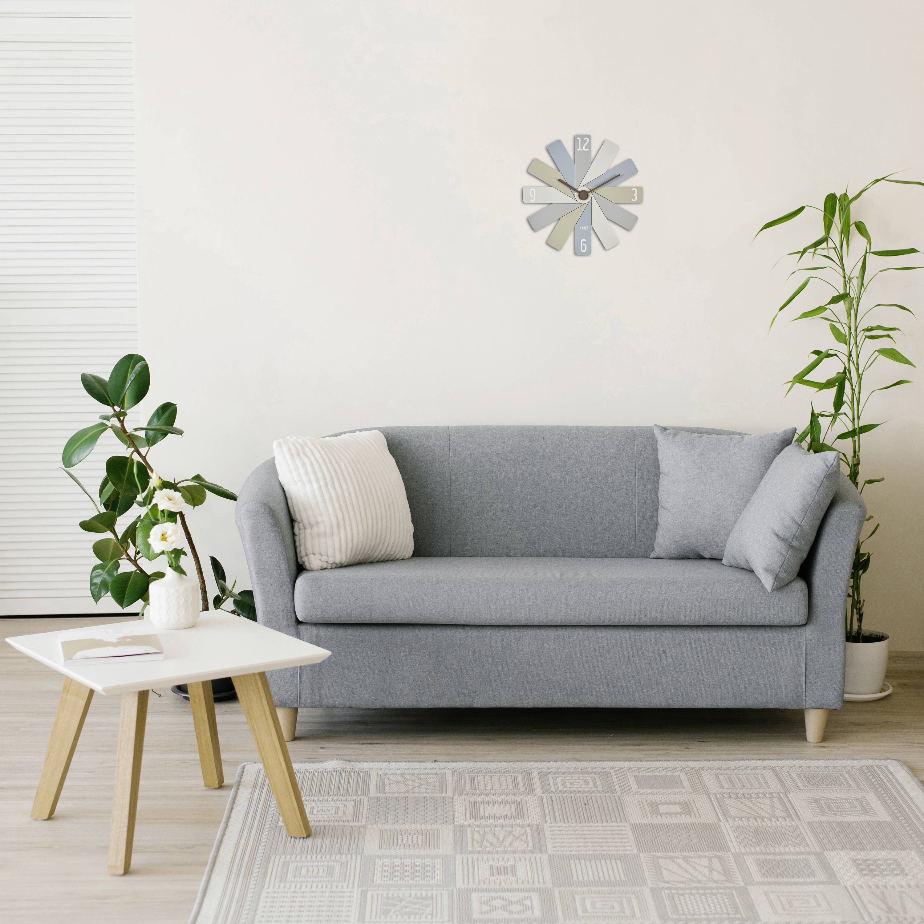 Helles Wohnzimmer mit grauem Sofa, Kissen, Pflanzen und rundem Tisch. Eine moderne Uhr hängt an der Wand.
