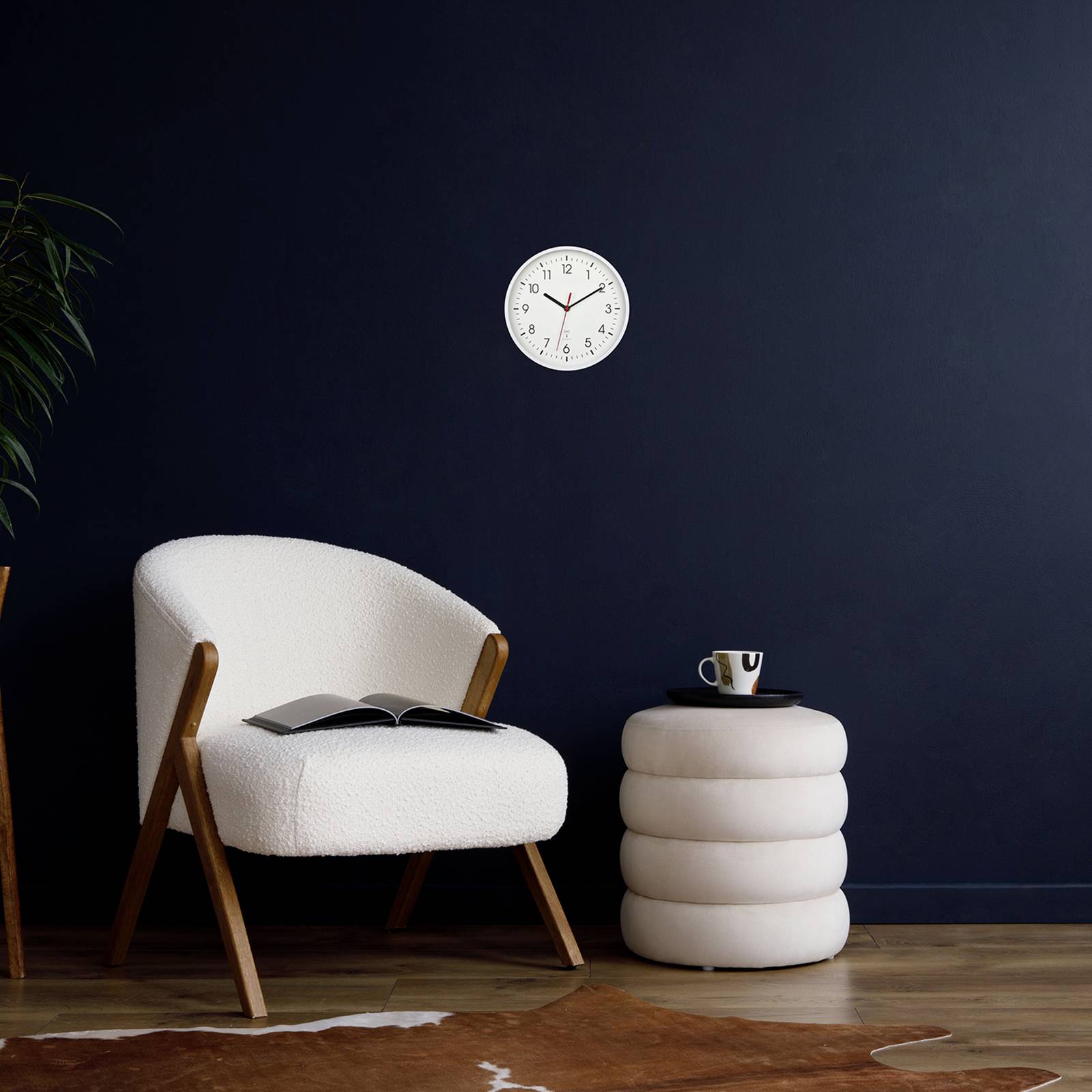 Ein beiger Sessel und ein beistellender Hocker mit Tasse vor einer dunkelblauen Wand. Eine Uhr zeigt 10:09 an.