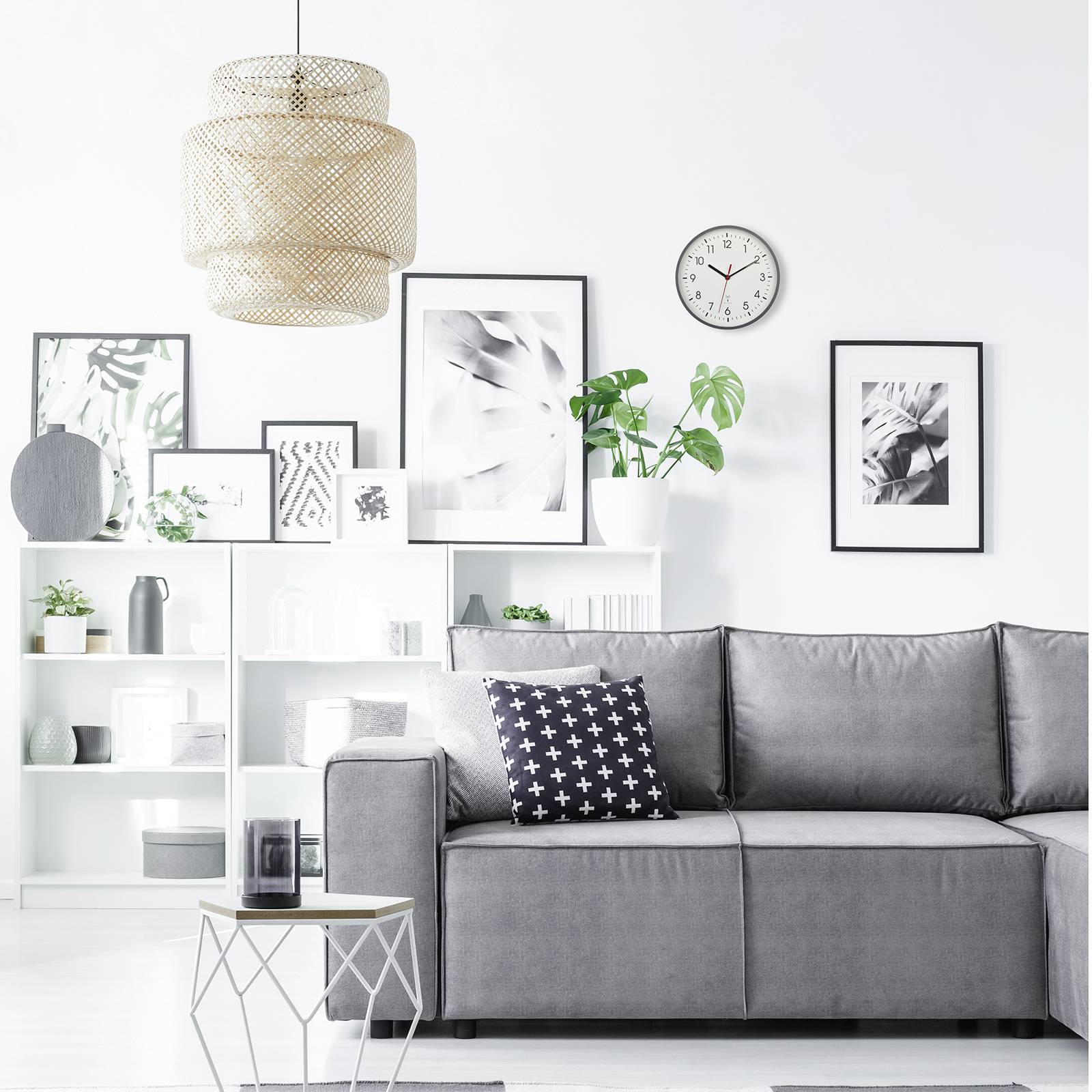 Ein modernes Wohnzimmer mit grauem Sofa, dekorativen Kissen, weißem Regal mit Pflanzen und Bilderrahmen an der weißen Wand.