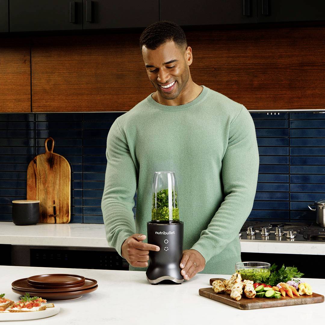 Ein Mann lächelt, während er einen Mixer mit grünem Smoothie-Inhalt auf einer Küchenarbeitsplatte hält. Im Hintergrund Küchenutensilien.
