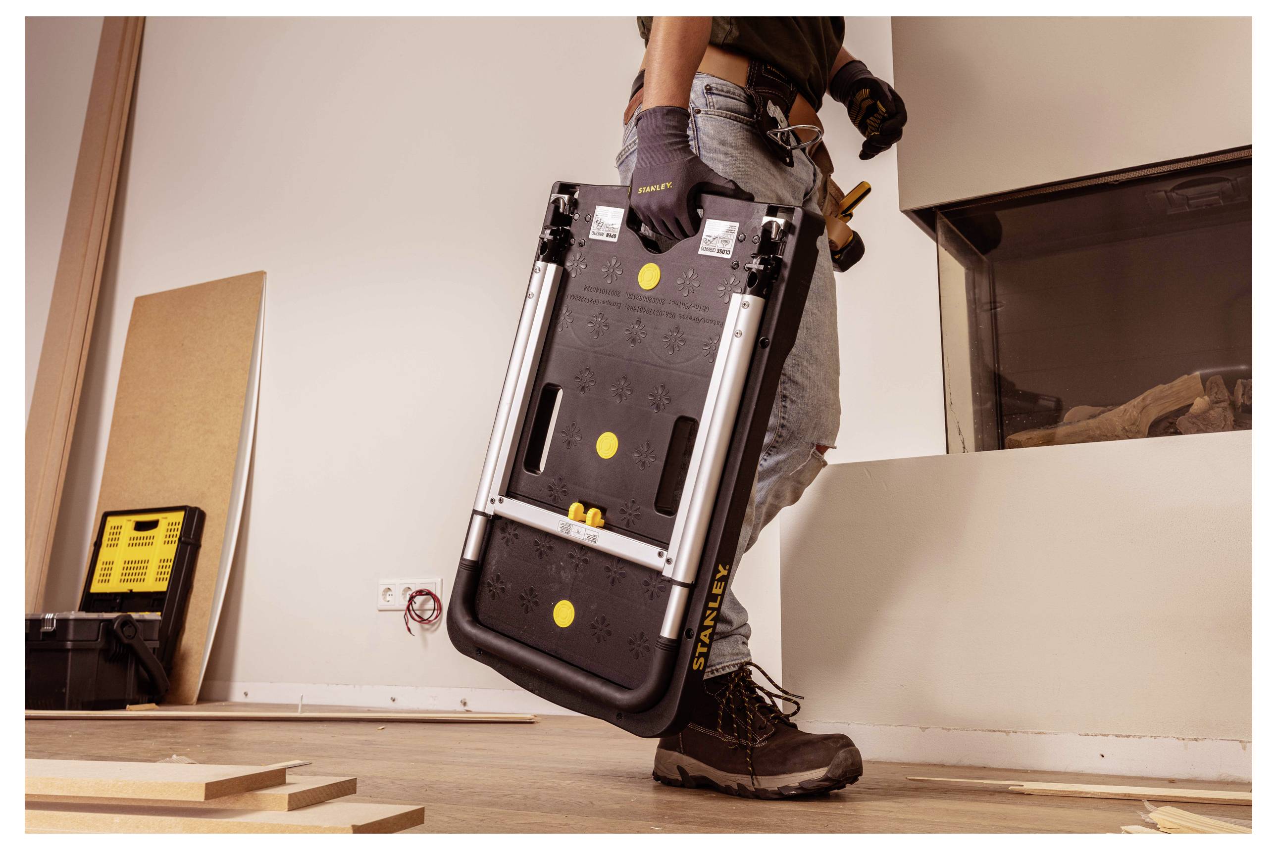 Eine Person hält eine zusammengeklappte, tragbare Werkbank in einer Werkstatt-Umgebung, trägt Handschuhe und Stiefel und deutet damit an, dass sie sich auf ein Projekt vorbereitet.