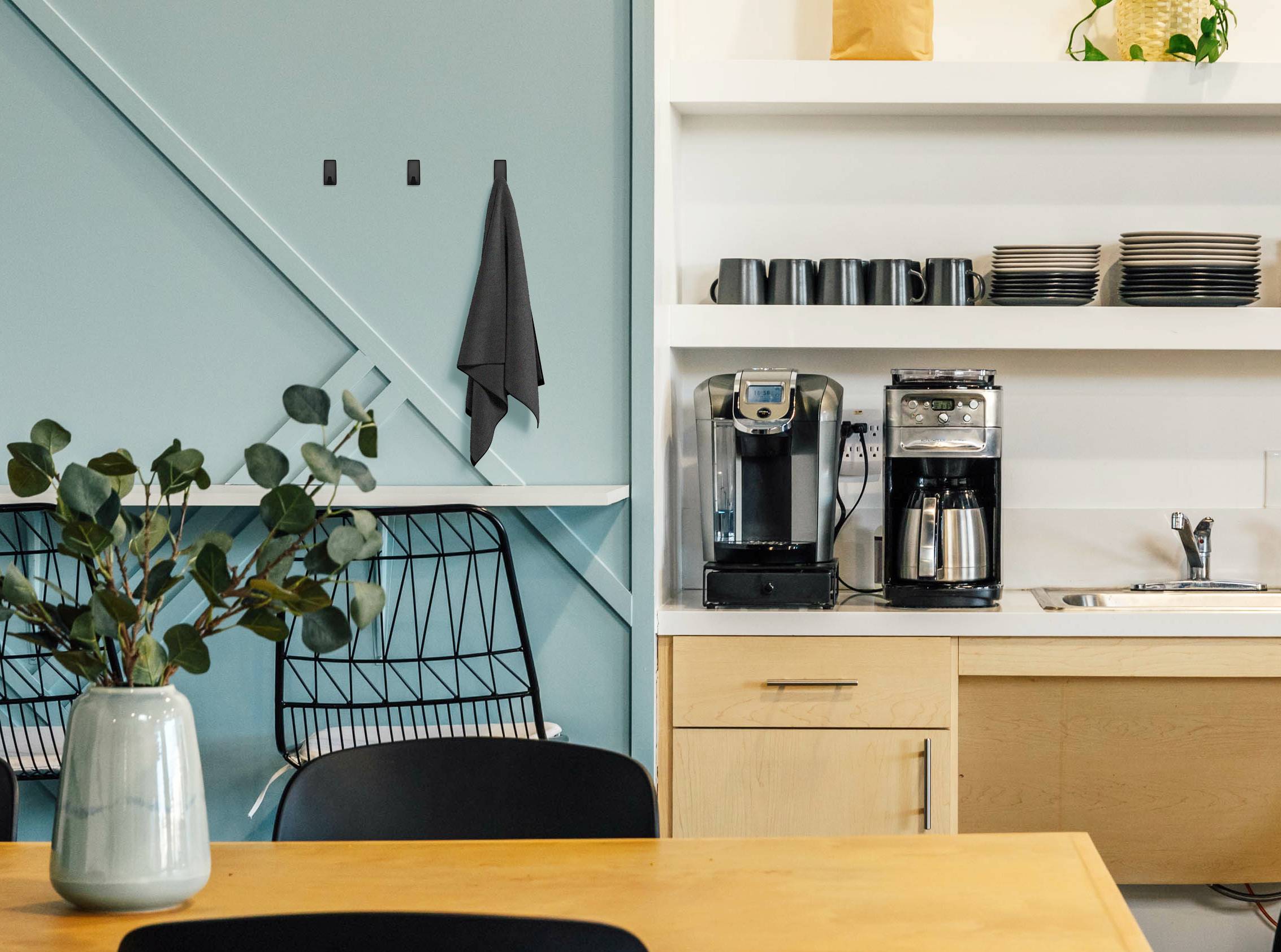 Moderner Kaffeeraum mit Kaffeemaschine, Tassen und Geschirrregal. Zwei Drahtstühle, Tisch mit Vase und Pflanzen. Wand in Pastellgrün.