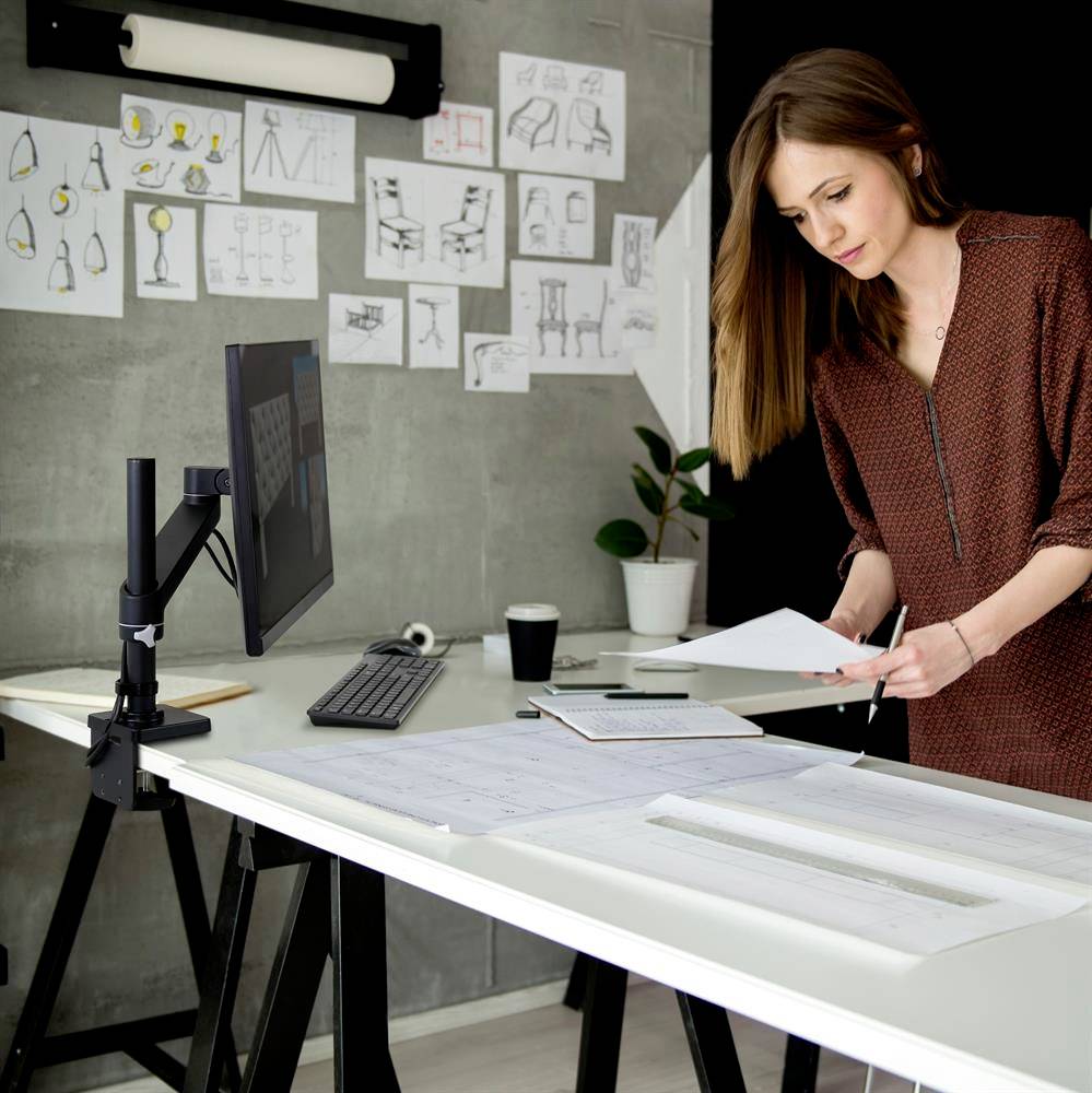 Eine Frau steht an einem Schreibtisch mit Monitor und überprüft Pläne. An der Wand hängen Zeichnungen und Entwürfe von Lampen und Möbeln.