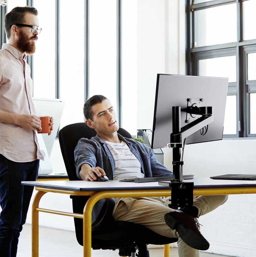 Zwei Männer im Büro, einer sitzt am Schreibtisch mit Computer, der andere steht und hält eine Tasse. Helle, moderne Umgebung.