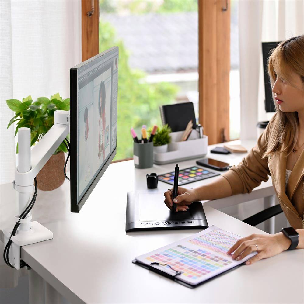 Frau arbeitet kreativ am Computer auf einem Grafiktablet, umgeben von Büromaterial. Fenster im Hintergrund lässt Tageslicht herein.