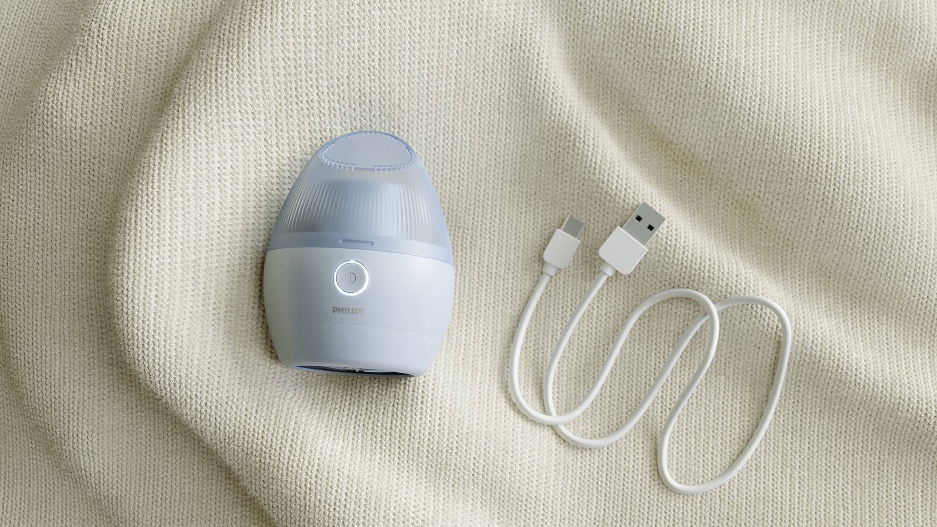 Ein kleines, rundes Gesichtsmassagegerät mit blauem Gehäuse und einem USB-Ladekabel liegt auf einem beigen Stoffhintergrund.