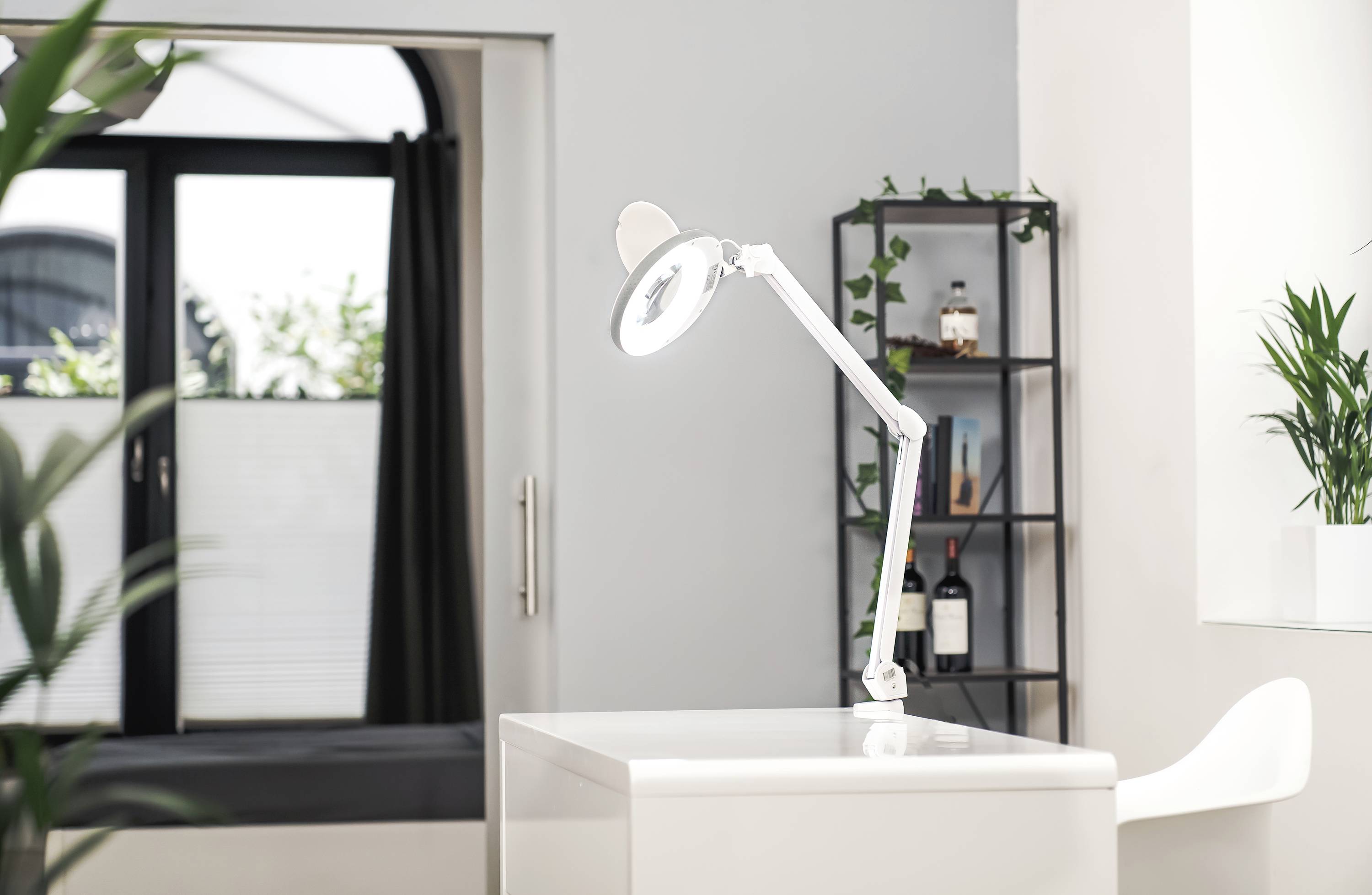 Ein Schreibtisch mit einer modernen Schreibtischlampe in einem hellen Raum, umgeben von Pflanzen. Ein Regal im Hintergrund mit Flaschen.