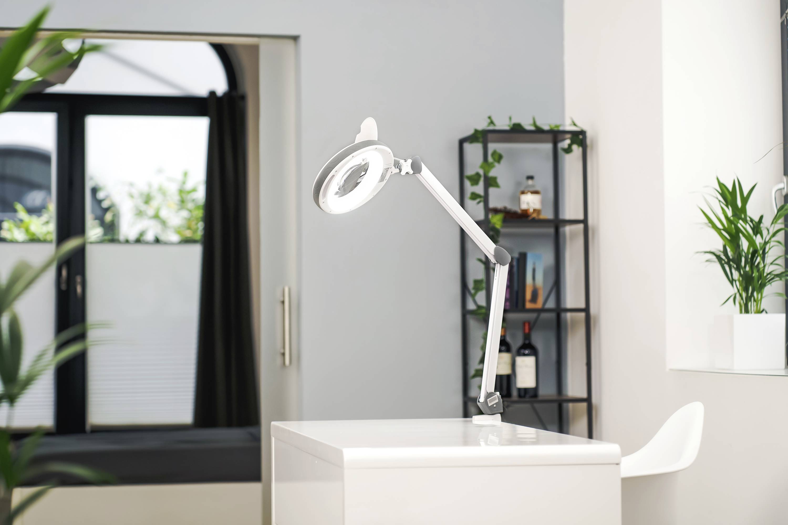 Ein heller, moderner Arbeitsbereich mit einem weißen Schreibtisch, einer verstellbaren Lampe und Pflanzen im Hintergrund.