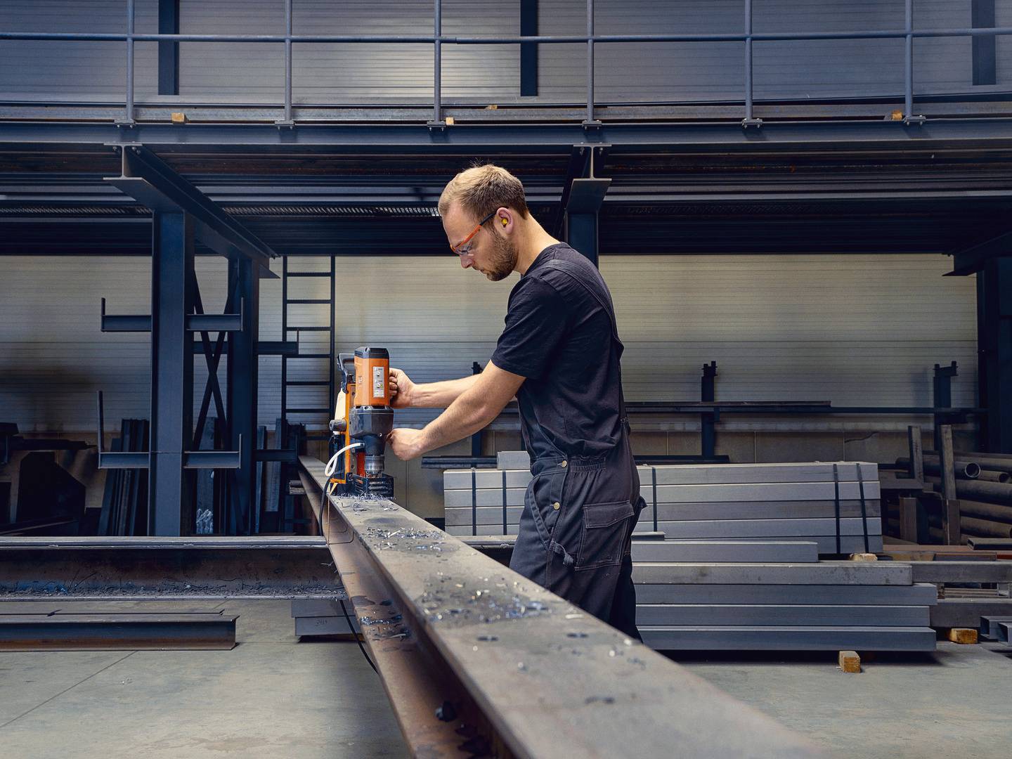 Ein Mann arbeitet in einer Werkstatt und benutzt eine Bohrmaschine, um ein langes Metallstück zu bearbeiten. Im Hintergrund sind Stahlträger zu sehen.