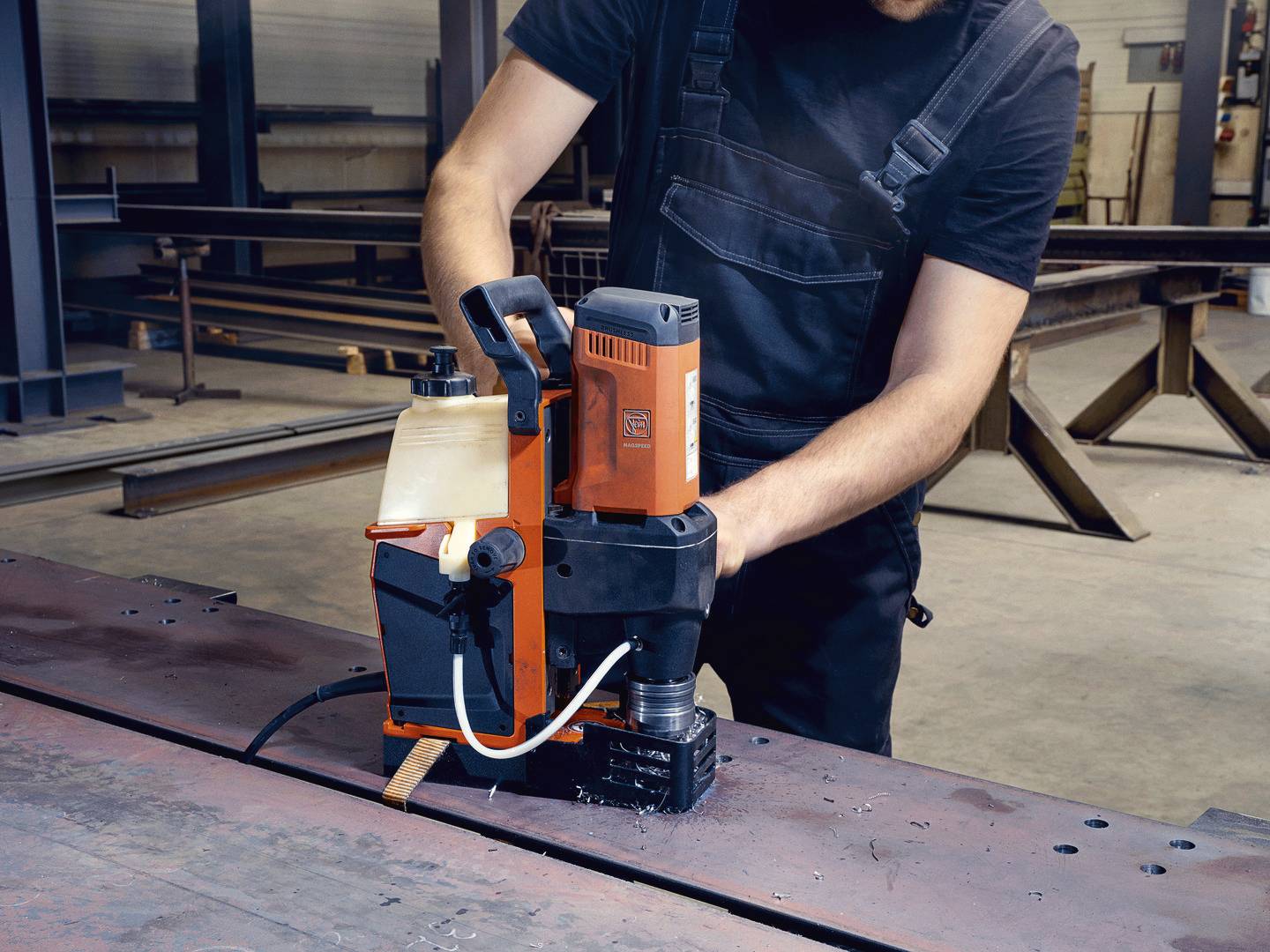 Eine Person in Arbeitskleidung bedient eine magnetische Bohrmaschine auf einer Metallplatte in einer Werkstattumgebung.
