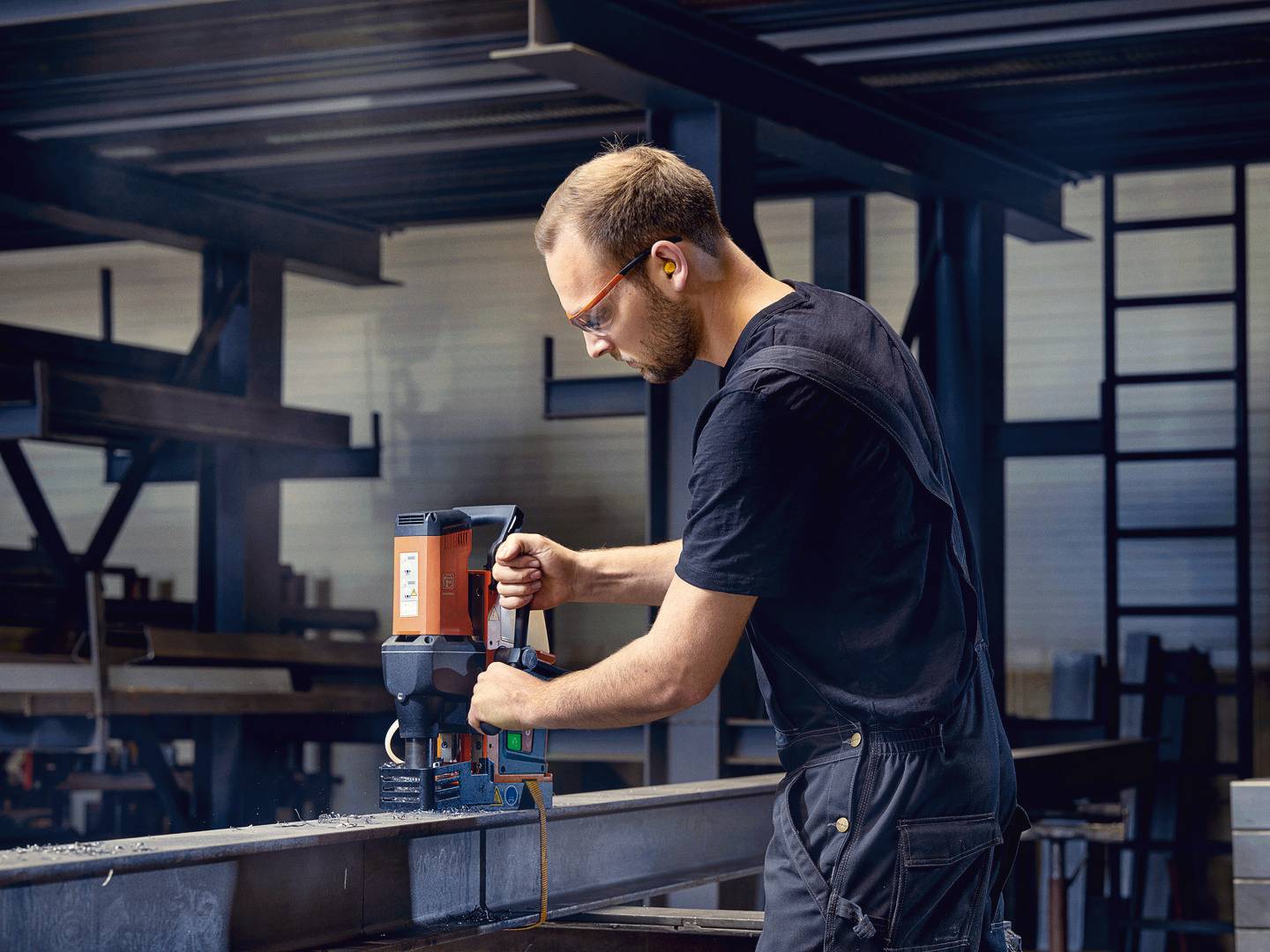 Ein Mann in Schutzbrille und Overall arbeitet in einer Werkhalle mit einem magnetischen Bohrer an einem Stahlträger.