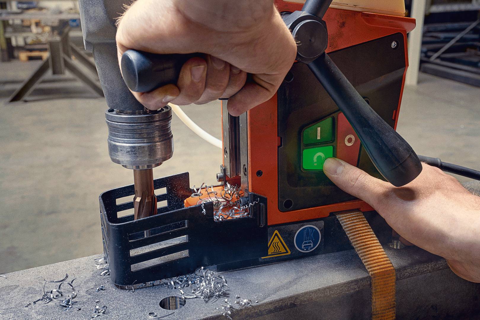 Eine Person bedient eine industrielle Bohrmaschine, um ein Metallstück zu bohren. Hände halten Bohrer und Maschine. Schaltflächen sind sichtbar.