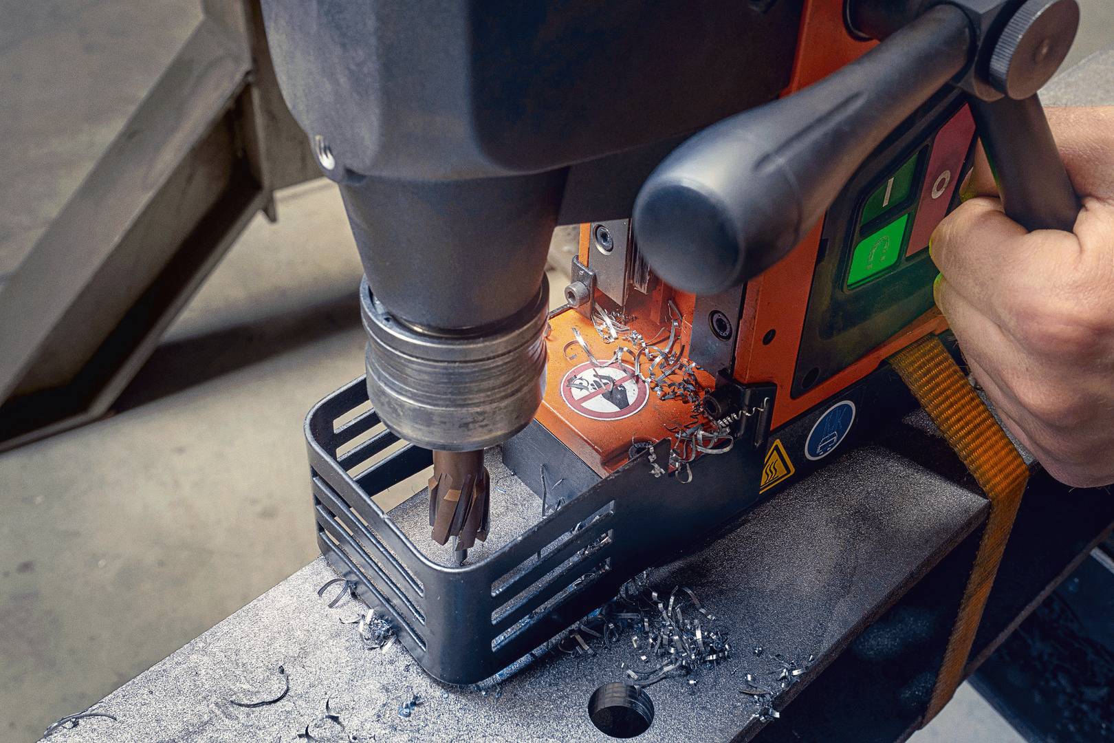 Eine Person bedient eine Bohrmaschine, die Metallspäne auf einen Metalltisch fallen lässt. Sicherheitszeichen auf der Maschine sichtbar.